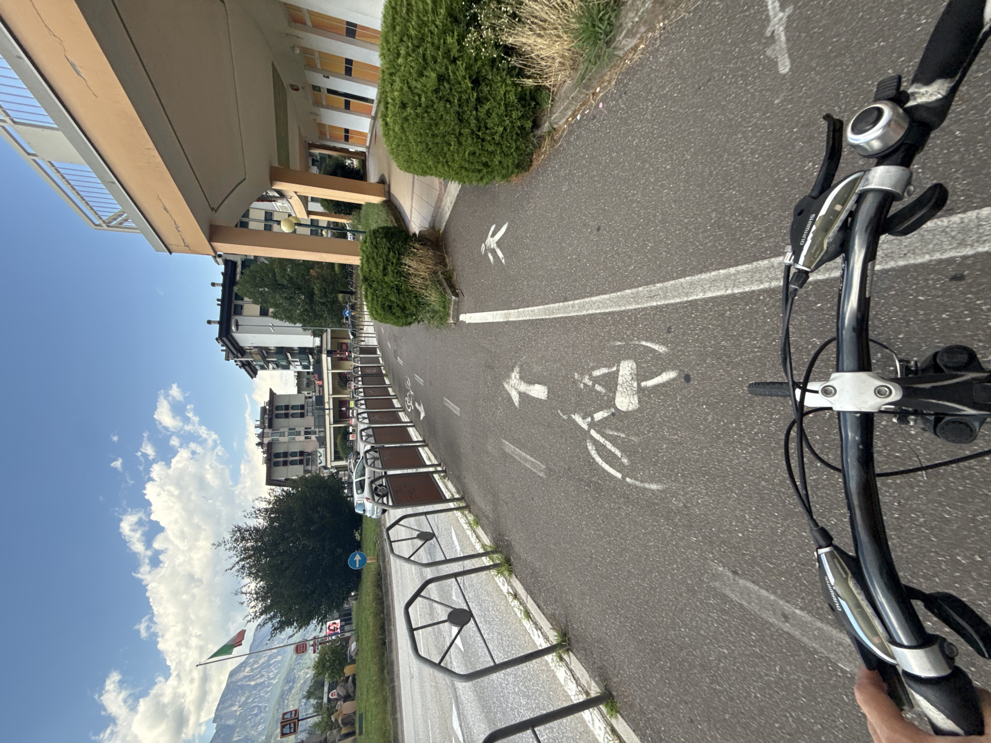 The image shows a first-person perspective of someone riding a bicycle on a dedicated bike path in Trento, Italy. The bike path is paved and marked with white lines, a bicycle symbol, and a pedestrian symbol. To the left of the bike path is a road with traffic and a metal barrier separating it from the bike path. In the background, there are buildings, trees, a mountain range, and the Italian flag. The weather appears to be sunny with some clouds in the sky. The bike's handlebars and brake levers are visible in the foreground, and a hand is on the left handlebar.