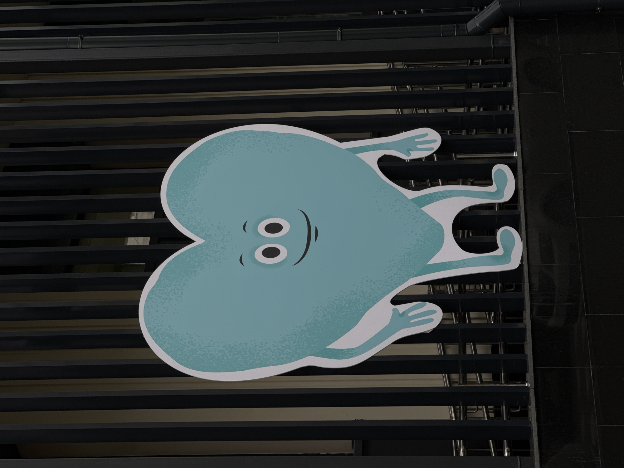 A light blue, heart-shaped cartoon character with wide eyes, a smiling mouth, and thin arms and legs is standing in front of a dark metal fence. The character has a textured, speckled appearance. The fence is composed of vertical slats with a gap between them, behind which is a blurred background with some light-colored elements. The bottom of the image shows a dark tiled surface. The scene is outdoors, with the character appearing to be a cutout or a prop. The location is not specified.