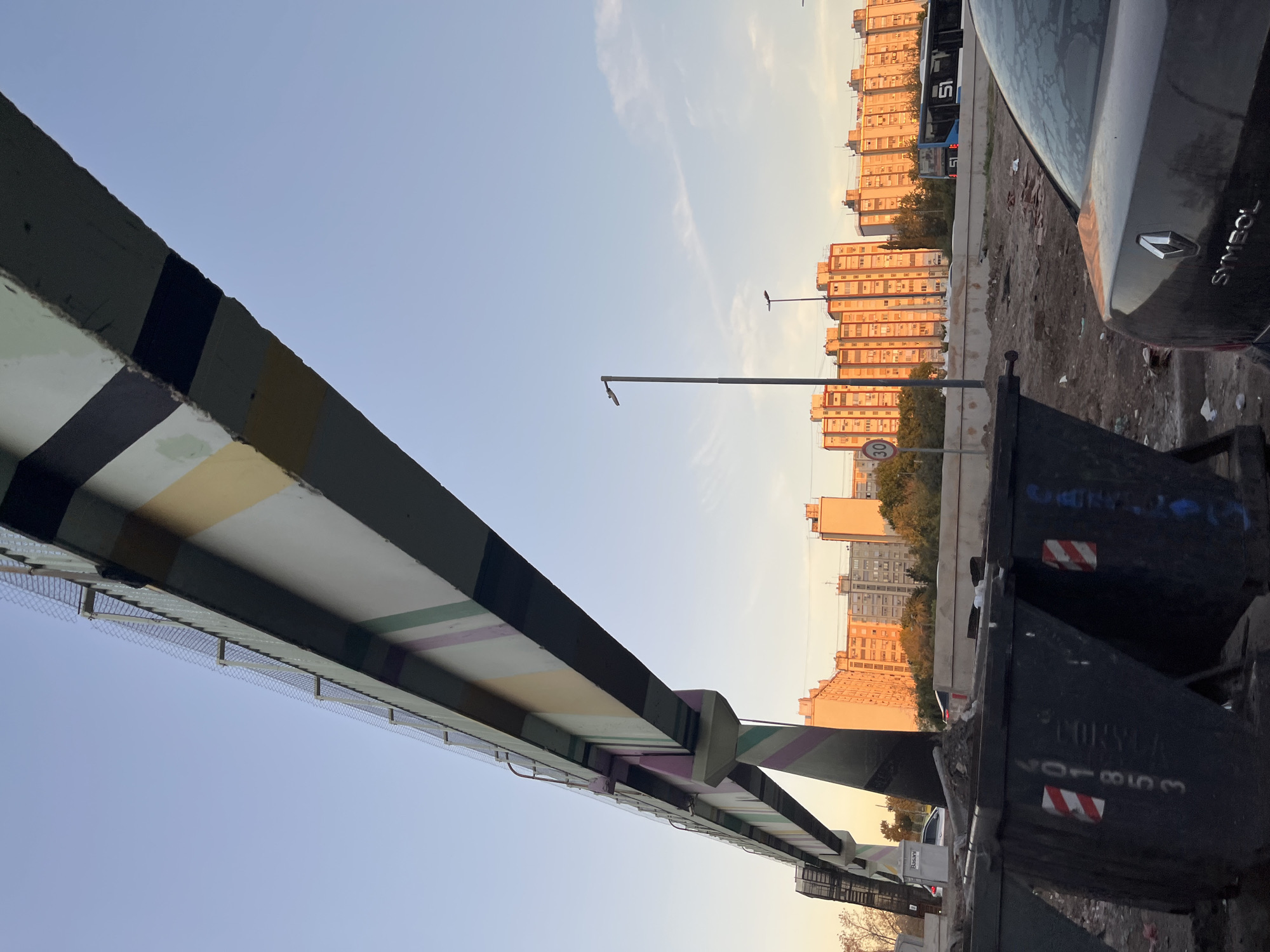 The image depicts an urban landscape during what appears to be the golden hour, either sunrise or sunset, characterized by warm, golden light illuminating the buildings. The perspective is from beneath a large concrete overpass or bridge, which spans across the upper left and center of the frame. This structure is painted with various horizontal stripes in black, dark grey, white, light yellow, and pastel green/purple, and features a protective wire mesh net running along its underside.
In the foreground, on the right, are two large, dark-colored industrial waste containers, each bearing red and white striped hazard markings. One of the bins has the text "CORYCA" and the number "201853" visible. Adjacent to these bins, a dark-colored car is parked, partially visible, with the word "SYMBOL" and the Renault logo clearly distinguishable on its trunk. The ground beneath the car and bins is an unpaved, uneven surface with debris.
Beyond the immediate foreground, a line of multi-story residential or commercial buildings stretches across the midground, their facades aglow with the warm sunlight. Scattered trees are visible in front of and between some of these buildings. Several street lights punctuate the scene, and one sign with the number "30" in a red circle, likely indicating a speed limit, is visible. A blue and white bus can be seen in the distance, passing by the buildings. The sky above is clear and light blue with a few faint wisps of clouds, suggesting fair weather. No people are visible in the scene. The overall setting is an active urban area, likely near a highway or a major road.