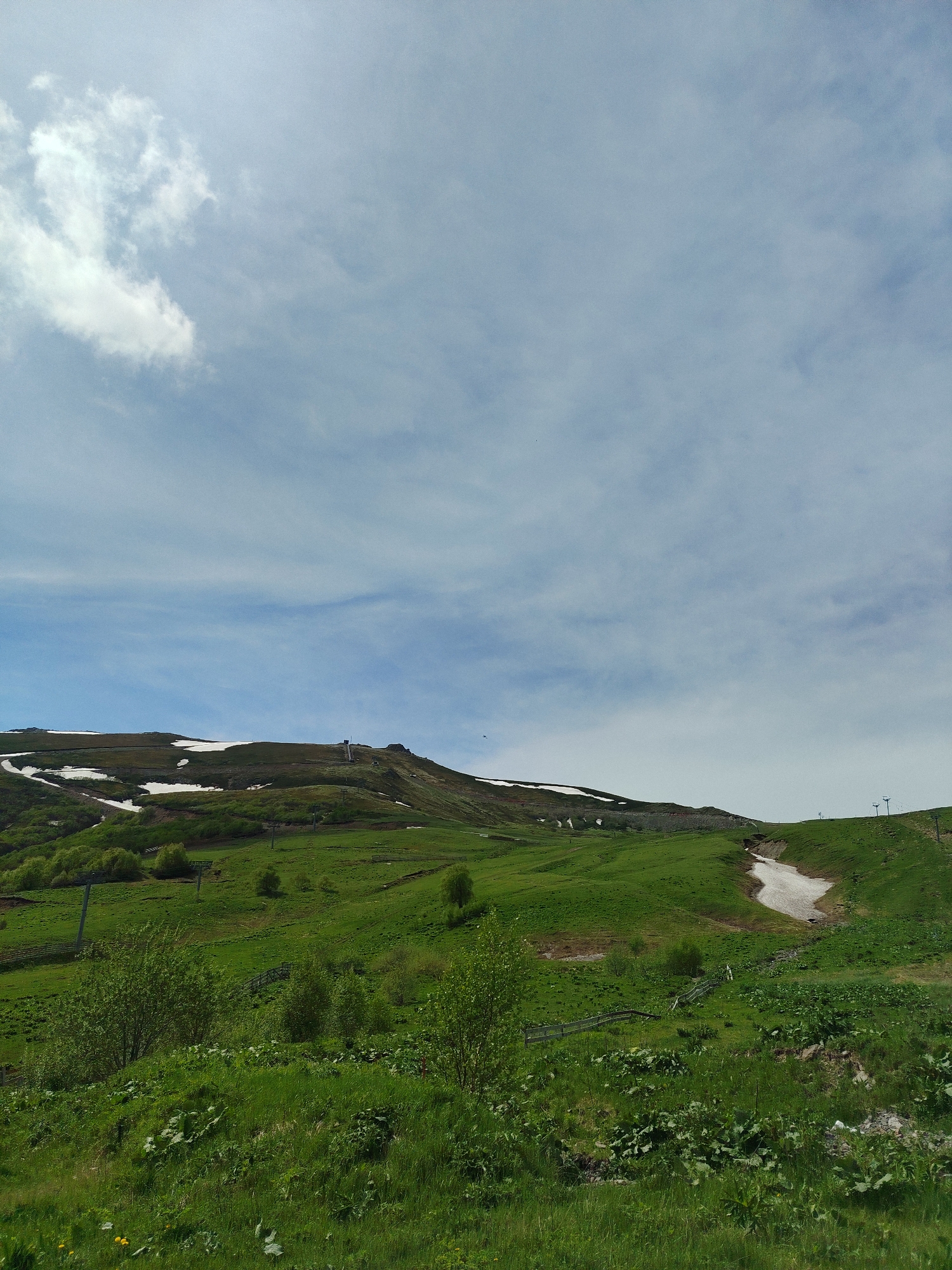 The image depicts a scenic view of a grassy mountainside with patches of snow. The sky is partly cloudy, with white clouds scattered against a pale blue background. In the mid-ground, a ski lift with poles and cables is visible, indicating a ski resort. The terrain is uneven and slopes upwards towards the peak, which is partially covered in snow. The foreground is dominated by lush green grass, with some bushes and smaller plants. The scene suggests a natural landscape in a mountainous region, possibly during the spring or early summer when snow still lingers on higher elevations. The location is identified as Bakuriani, Georgia, a well-known mountain resort. There are no people or animals visible in the image. The overall atmosphere is serene and picturesque.