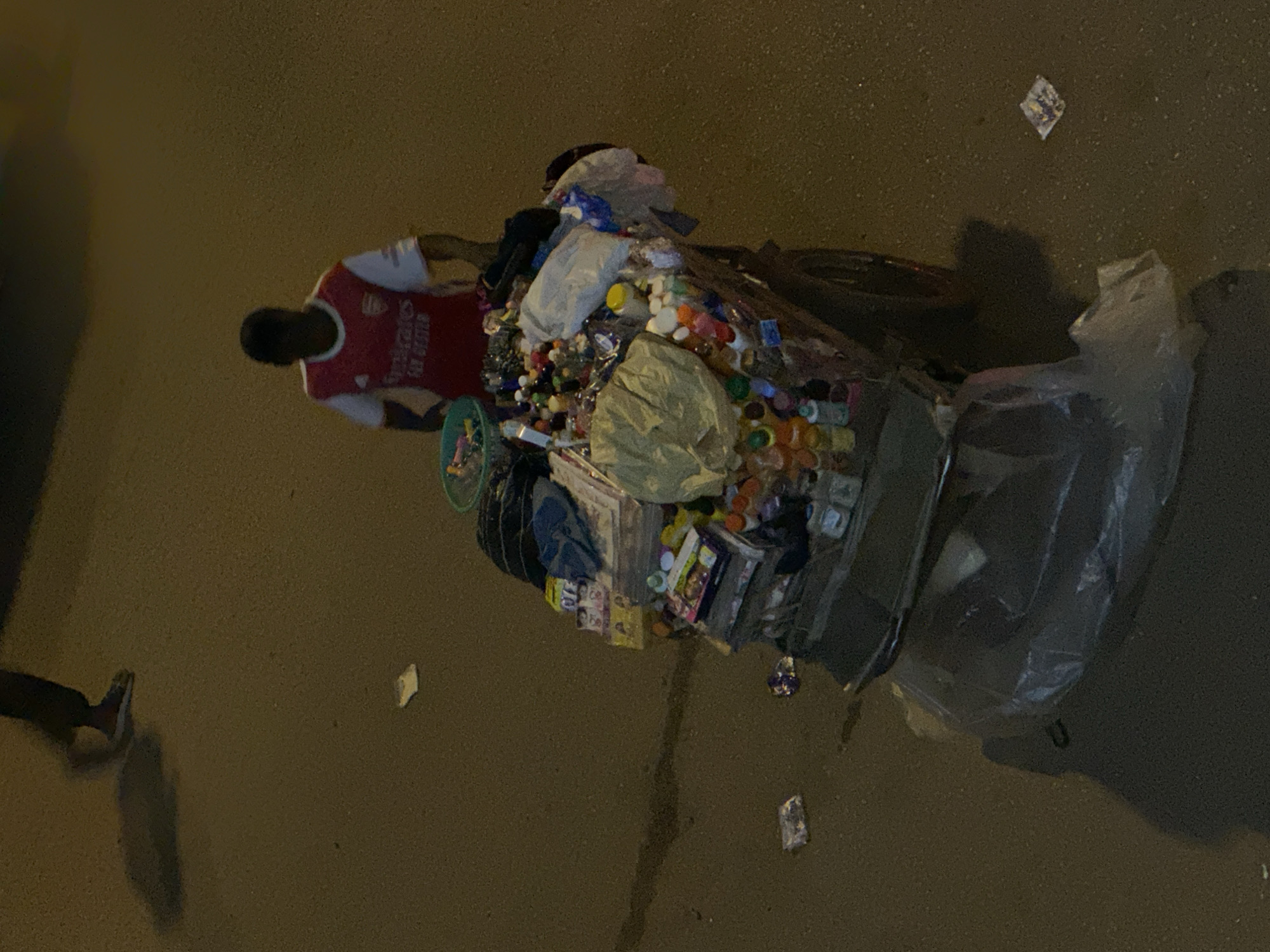An elevated, full shot captures a street vendor in Bukuru, Nigeria, standing beside a cart laden with various goods. The vendor, wearing a red and white Arsenal football jersey, is partially obscured by the cart. The cart is filled with an assortment of items, including bottled drinks, packaged snacks, and what appear to be personal care products. A green plastic bowl sits atop the cart, containing smaller items. The ground around the cart is a mixture of dirt and asphalt, with scattered pieces of litter visible. A shadow stretches across the ground from an unseen person in the upper left of the frame, and the overall lighting suggests it is evening or nighttime.