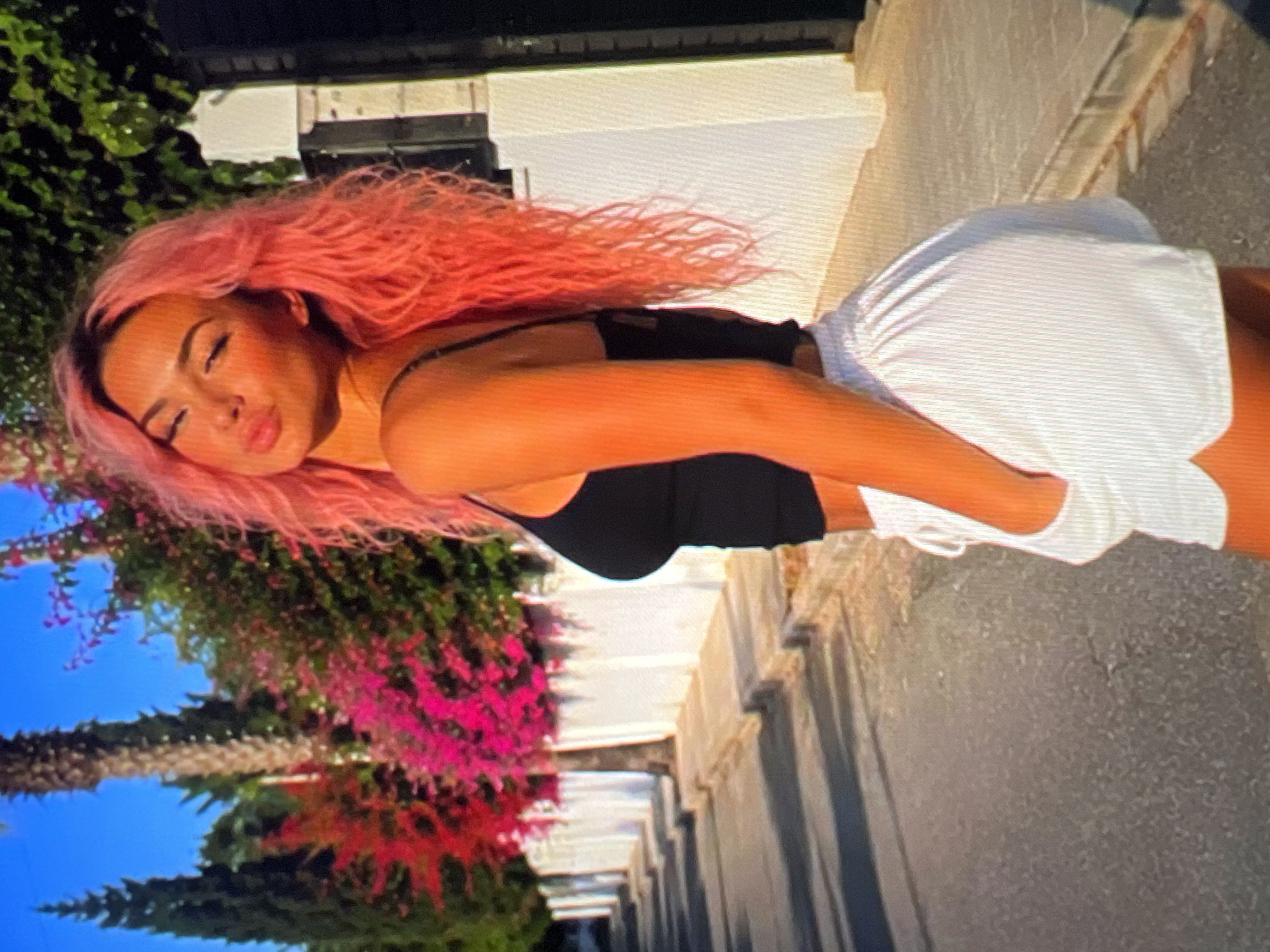 A woman with bright pink, wavy hair stands in profile on a paved road, facing left. She is wearing a black tank top and white shorts, with her left hand tucked into the pocket of her shorts. Her eyes are closed and her lips are pursed as if she is blowing a kiss. Behind her, a white wall lines the street, bordered by a sidewalk. A line of trees with vibrant pink and red flowers grows along the wall. The sun casts long shadows across the road.