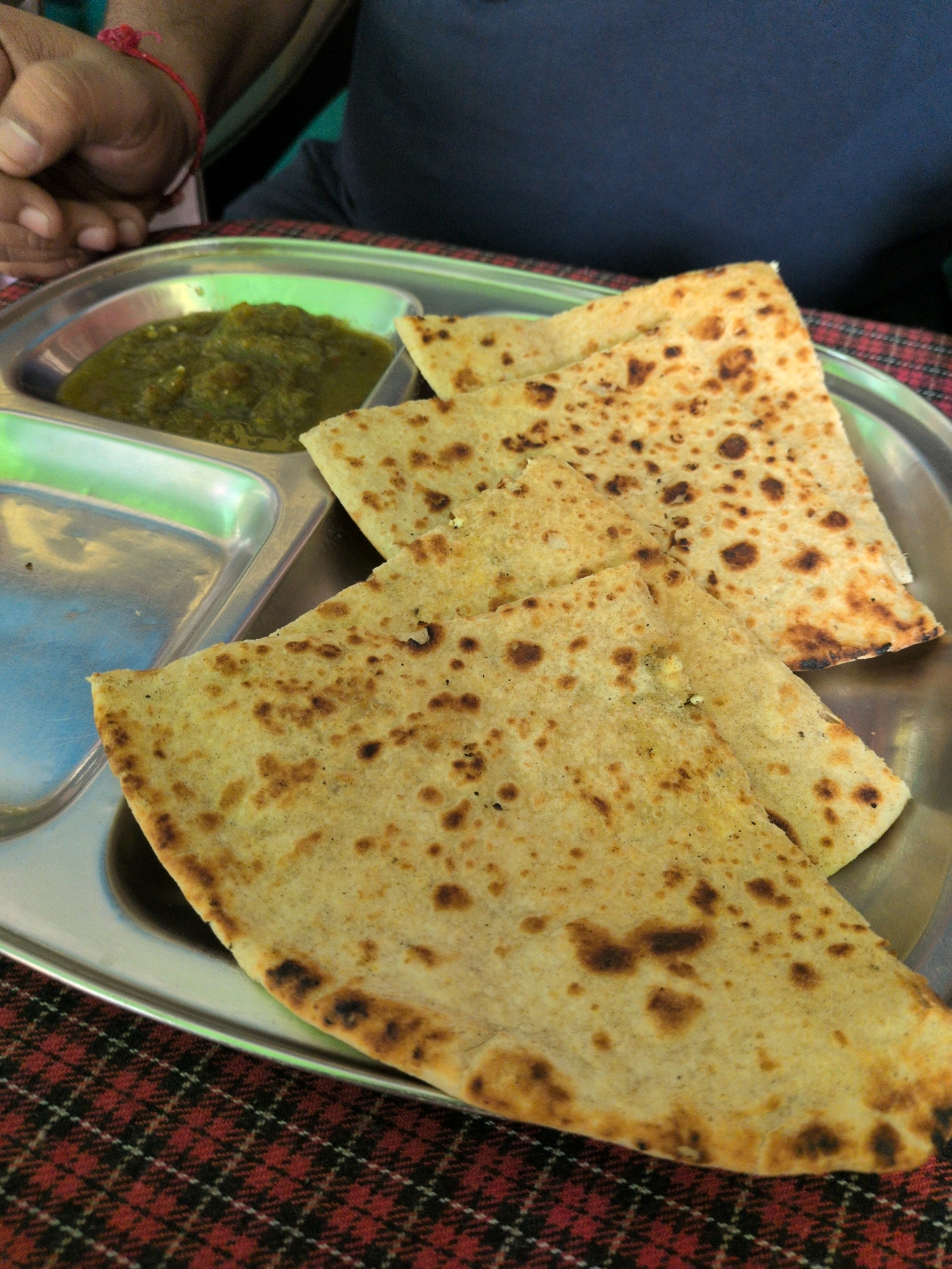 A close-up shot shows a metal tray with Indian food on a red and black checkered cloth. The tray contains a green chutney in a small compartment and several pieces of paratha, a type of flatbread, filling the majority of the frame. The paratha are golden brown with darker, toasted spots. A person's hands, wearing a red thread bracelet, are visible in the upper left corner, suggesting a person is about to eat the food. The image is likely taken in Vashisht, India, a town known for its hot springs and spiritual retreats.