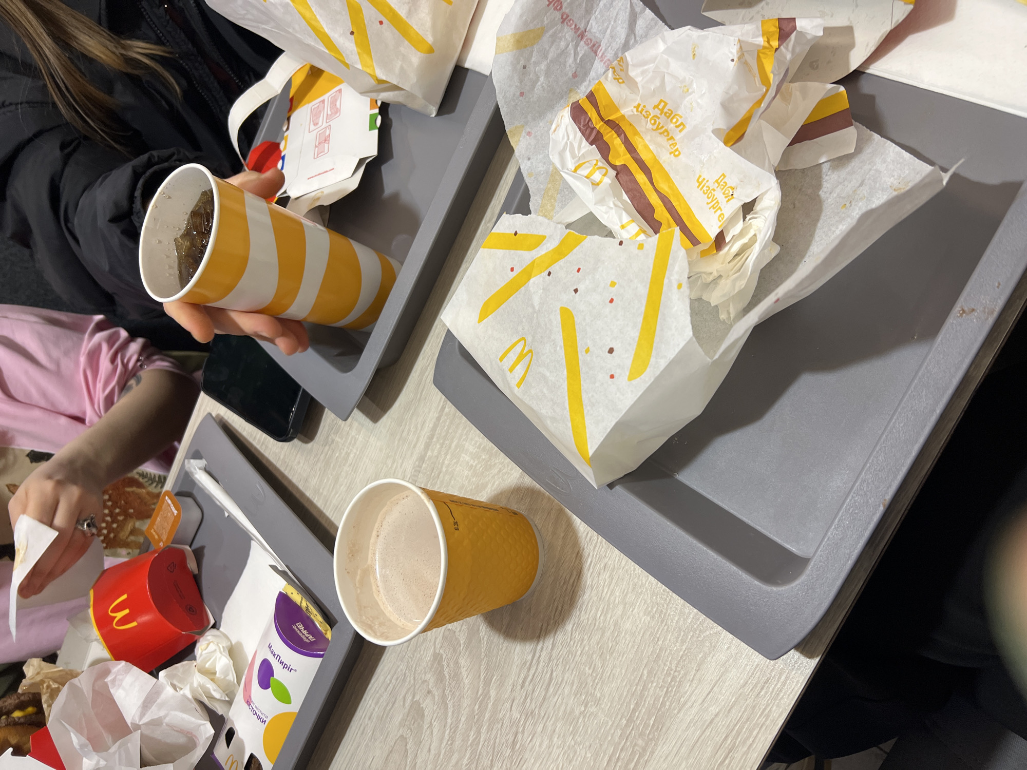 The image shows a table at a McDonald's restaurant in Odesa, Ukraine. There are two gray trays with McDonald's packaging. One tray holds an unfinished McDonald's Coca-Cola cup, burger wrappers, and a drink holder. The other tray has a McDonald's McFlurry container, a fry container, and a burger wrapper. A person with a black jacket and a pink shirt is sitting at the table, eating a burger. There is also a smartphone on the table. The table has a light wood-grain surface.