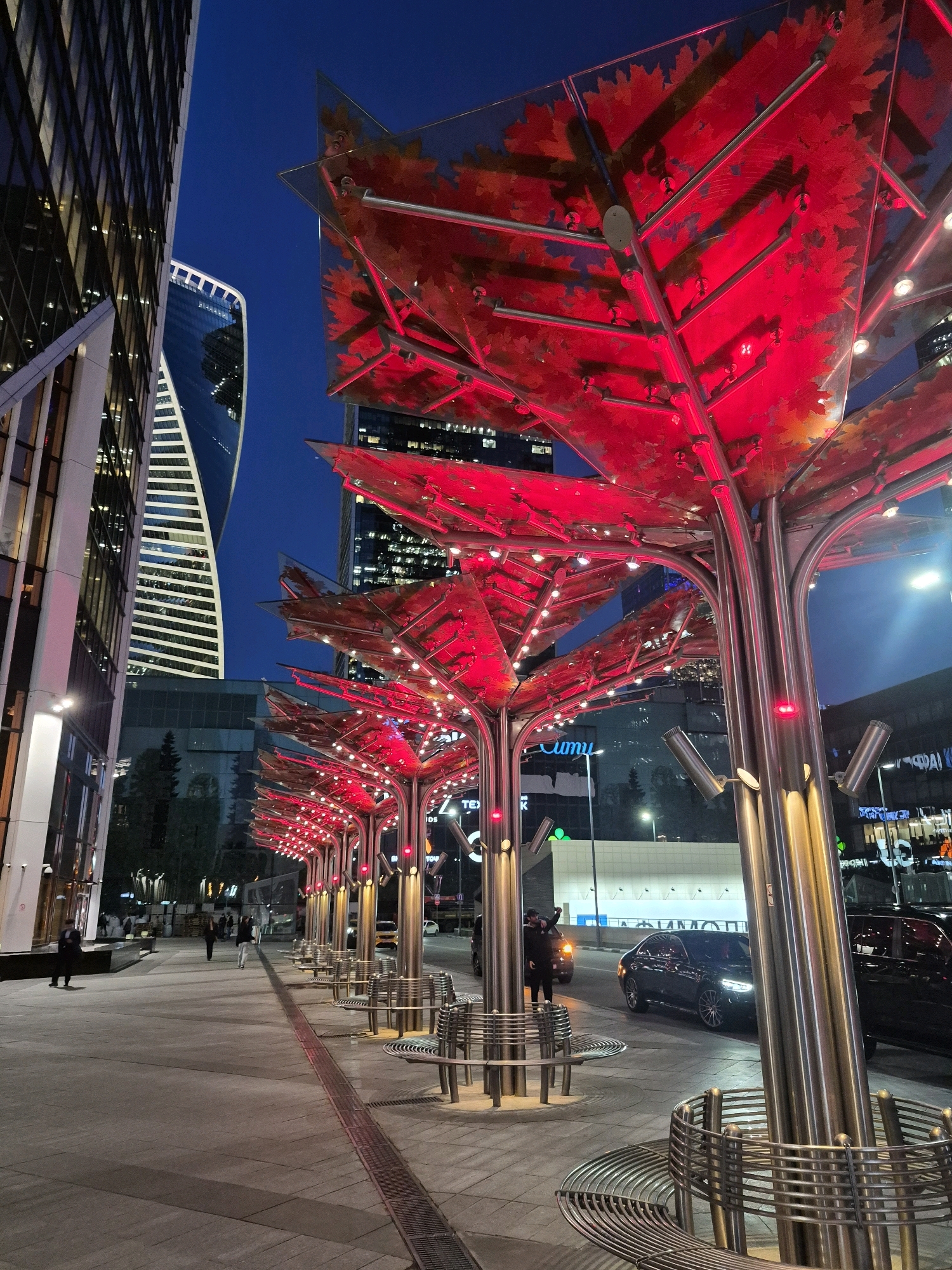 The image depicts a nighttime scene in a modern urban environment, likely a city square or pedestrian area. In the foreground, a series of decorative structures resembling stylized trees with bright red, leaf-shaped canopies illuminated from within are lined up along a paved walkway. These structures have metallic trunks and branches, and are adorned with numerous small lights. Metallic benches with a circular design are placed around the base of these decorative trees.
To the left, a tall, modern glass-and-steel building with a distinctive angled facade dominates the background. Further back, a curved skyscraper with horizontal window bands is visible. Other contemporary buildings with illuminated windows can be seen in the distance.
The walkway is made of concrete slabs with a red-lined strip running down its center. Several people are walking along the walkway. On the right side of the frame, a dark-colored sedan is parked on a street adjacent to the walkway, and another dark vehicle is partially visible behind it. The sky is a deep blue, indicating it is twilight or night. The overall impression is one of a well-lit, contemporary urban setting designed for public use.