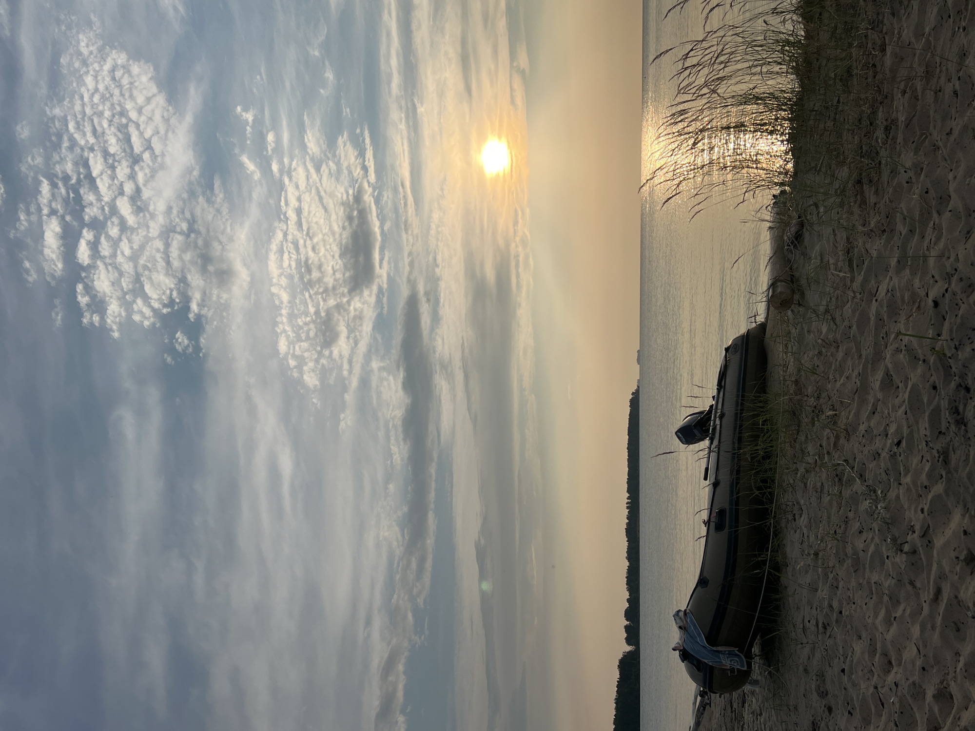 This image depicts a serene natural scene on a sandy beach, likely in Borovoe, Russia. The setting is a tranquil lake or sea meeting a sandy shore lined with tall, wispy grasses. In the foreground, an inflatable boat rests on the sand, with a towel draped over its side. The boat appears to be unoccupied.
The time of day is clearly late afternoon or early evening, as evidenced by the golden, diffused sunlight that bathes the scene. The sun is low on the horizon, its rays glinting off the water and casting long shadows. The sky is a canvas of soft, muted colors, with wispy clouds scattered across the blue expanse, adding texture and depth to the celestial view. The weather appears calm and pleasant, conducive to a peaceful moment by the water.
The overall mood is one of tranquility and natural beauty. The composition, with the vastness of the water and sky contrasted with the grounding presence of the sand and boat, evokes a sense of calm and relaxation. The dark silhouette of trees along the distant shoreline suggests a wooded area bordering the water body. There is no visible text in the image.