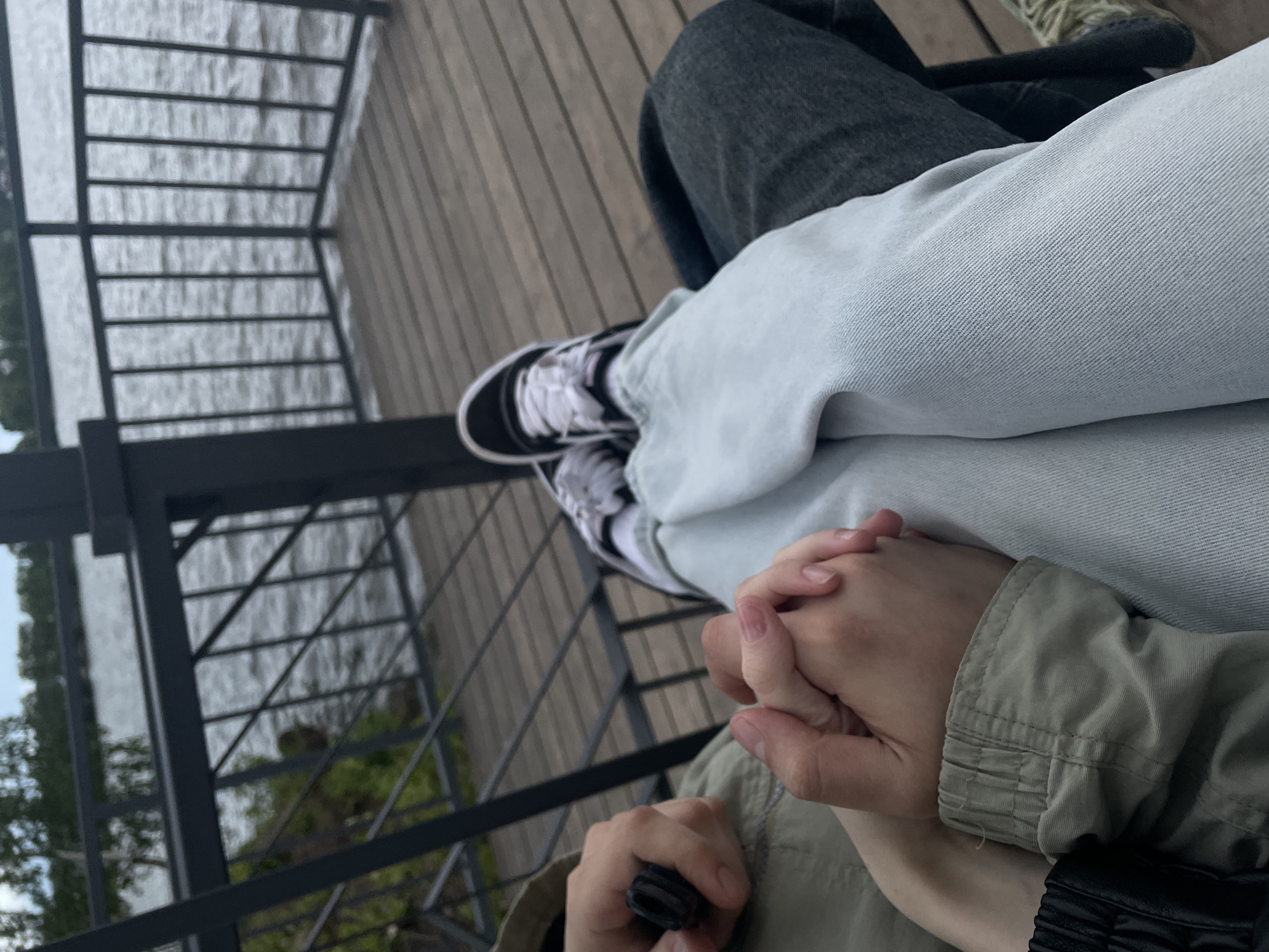 The image depicts a first-person, downward-angle perspective of two individuals on a wooden deck overlooking a body of water in Votkinsk, Russia. In the foreground, two hands are clasped together; one hand displays light pink manicured fingernails, while the other appears larger and less manicured. To the left, a person's right hand holds a small, black, cylindrical object. The person on the left wears an olive green or khaki-colored jacket with a ribbed cuff visible. The person on the right wears light-wash blue jeans and black high-top sneakers with white laces and soles, with their left leg crossed over a dark grey or black garment.
The wooden deck, composed of horizontal planks, extends into the mid-ground, where it is bordered by dark grey metal railings. These railings feature both vertical bars and diagonal cross-bracing in different sections, separating the deck from the water. Beyond the railings, a large expanse of water, likely the Votkinsk Pond, stretches into the background, showing ripples on its surface. The distant shore is lined with green trees and foliage, beneath a light sky. The scene suggests a calm outdoor setting on a designated viewing platform or pier.
