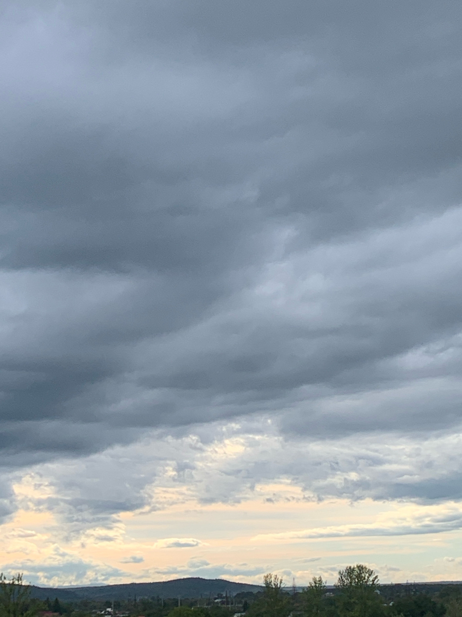 The image captures a panoramic view of the sky and the landscape of Kolomyia, Ukraine, under a thick, overcast sky. The clouds are predominantly dark gray, with some lighter patches suggesting breaks or thinner cloud layers. The bottom of the image reveals a horizon line with rolling hills or mountains forming a dark silhouette against a pale, hazy sky. Below the hills, a scattering of buildings and trees are visible, indicating a semi-urban or rural setting. The overall atmosphere is somber, possibly indicative of impending rain or a cloudy day. There are no people or distinct objects or actions identifiable in the scene.