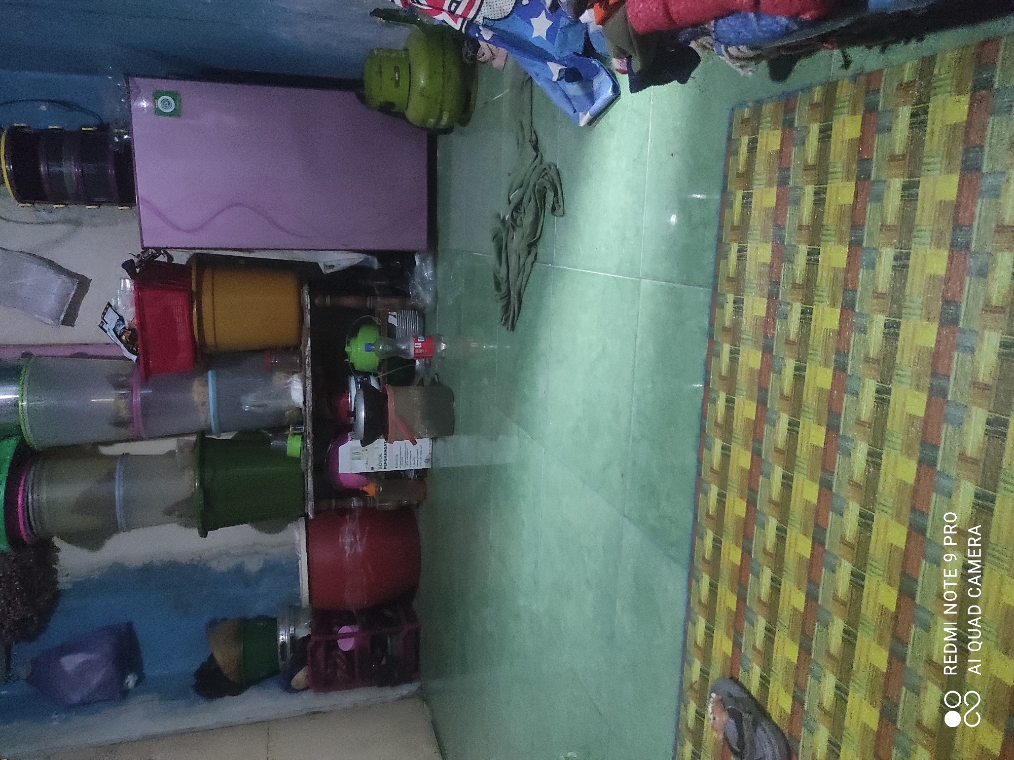 The image shows an interior scene in what appears to be a cluttered room, possibly a kitchen or storage area. The floor is tiled in a pale green color. In the foreground, a woven mat with a geometric pattern of yellow, red, and brown rectangular shapes is visible. A single sandal is placed on the mat.
Along the back wall, several items are stacked. On the left, there are various plastic containers and buckets in different colors, including blue, green, and red. A large red plastic drum is also present. To the right, a small, light pink refrigerator stands against the wall. Above the refrigerator, a large, dark purple water jug with black bands is visible. A green gas cylinder is situated next to the refrigerator.
Further to the right, a pile of fabric, primarily blue with white stars and red accents, is arranged on the floor. Scattered on the floor are a few more items, including a green t-shirt or cloth, a bottle of water, and a green gas cylinder.
The overall impression is one of domestic storage and everyday living. No individuals are clearly visible in the image. The environment suggests a functional space rather than a decorative one. The location is described as Sakra, Indonesia, but there are no specific visual cues in the image to confirm or deny this.