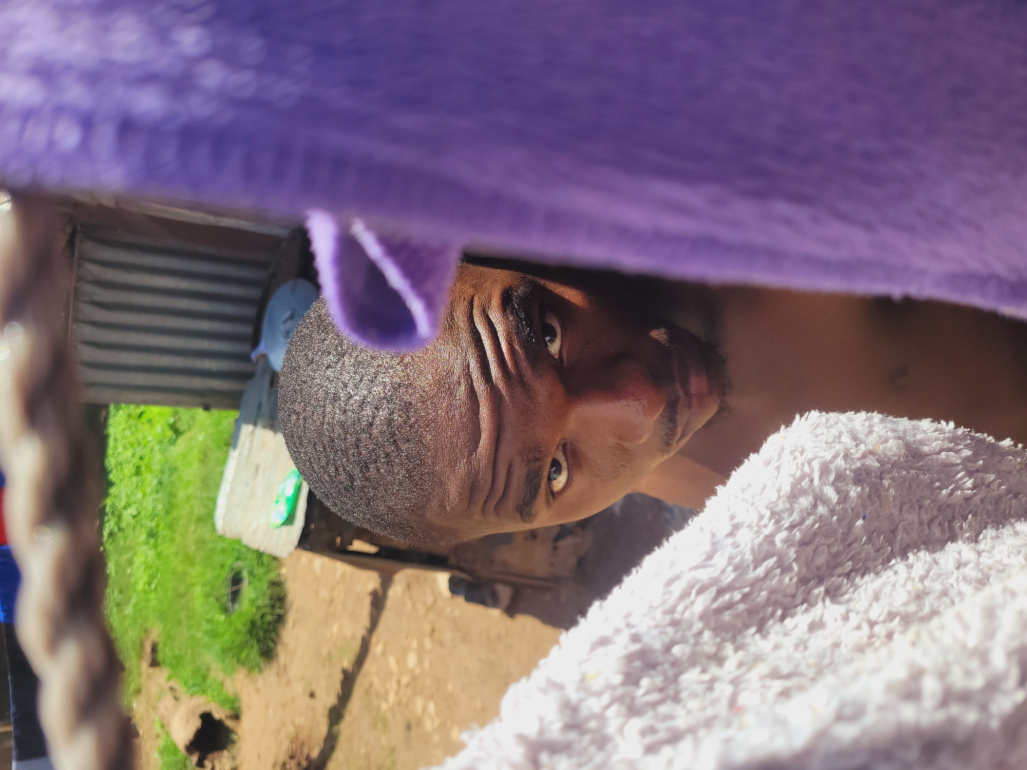 The image captures a close-up, low-angle portrait of a dark-skinned man in an outdoor setting in Bukuru, Nigeria. The man, who appears shirtless, looks directly into the camera with a neutral expression. He has short, dark hair and a short goatee. Visible wrinkles are present on his forehead.
The shot is framed by two soft, fluffy fabrics: a blurry purple one extends across the top of the image, and a light-colored (possibly off-white or cream) one is partially visible on the bottom right, both seemingly close to the camera lens.
In the background, behind the man, corrugated metal sheeting suggests a wall or structure. Beyond this, lush green grass and earthy, brown ground are visible. Bright sunlight illuminates the scene, creating clear highlights and shadows, indicating daytime. A dark circular feature is embedded in the grass, and a light-colored block with a green object rests on the ground. No visible text is present in the image.