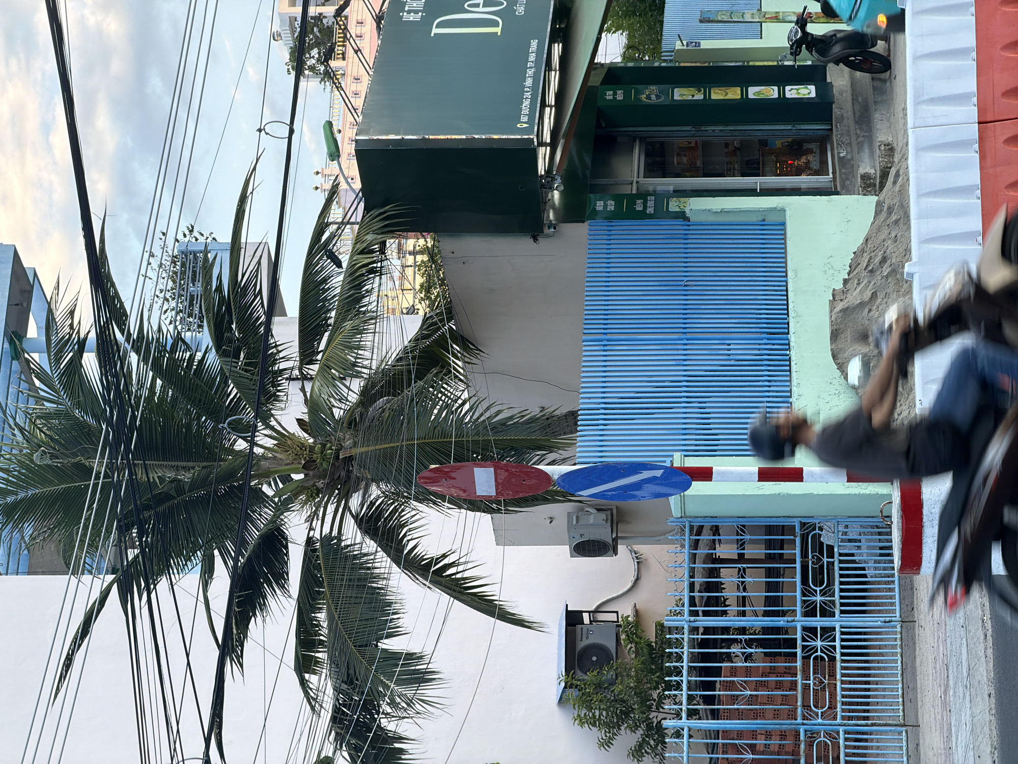 A man wearing a black helmet and a dark t-shirt is riding a motorcycle down a street in Bac Nha Trang Ward, Vietnam. He is in motion, and the motorcycle is blurred. In the background, there is a tall palm tree with a white building behind it. A blue fence with a red and white striped pole and traffic signs is visible. To the right, a green sign for "De" is displayed on the side of a building, with text below it in Vietnamese. A black scooter is parked in front of the building. The sky is overcast with some clouds.
