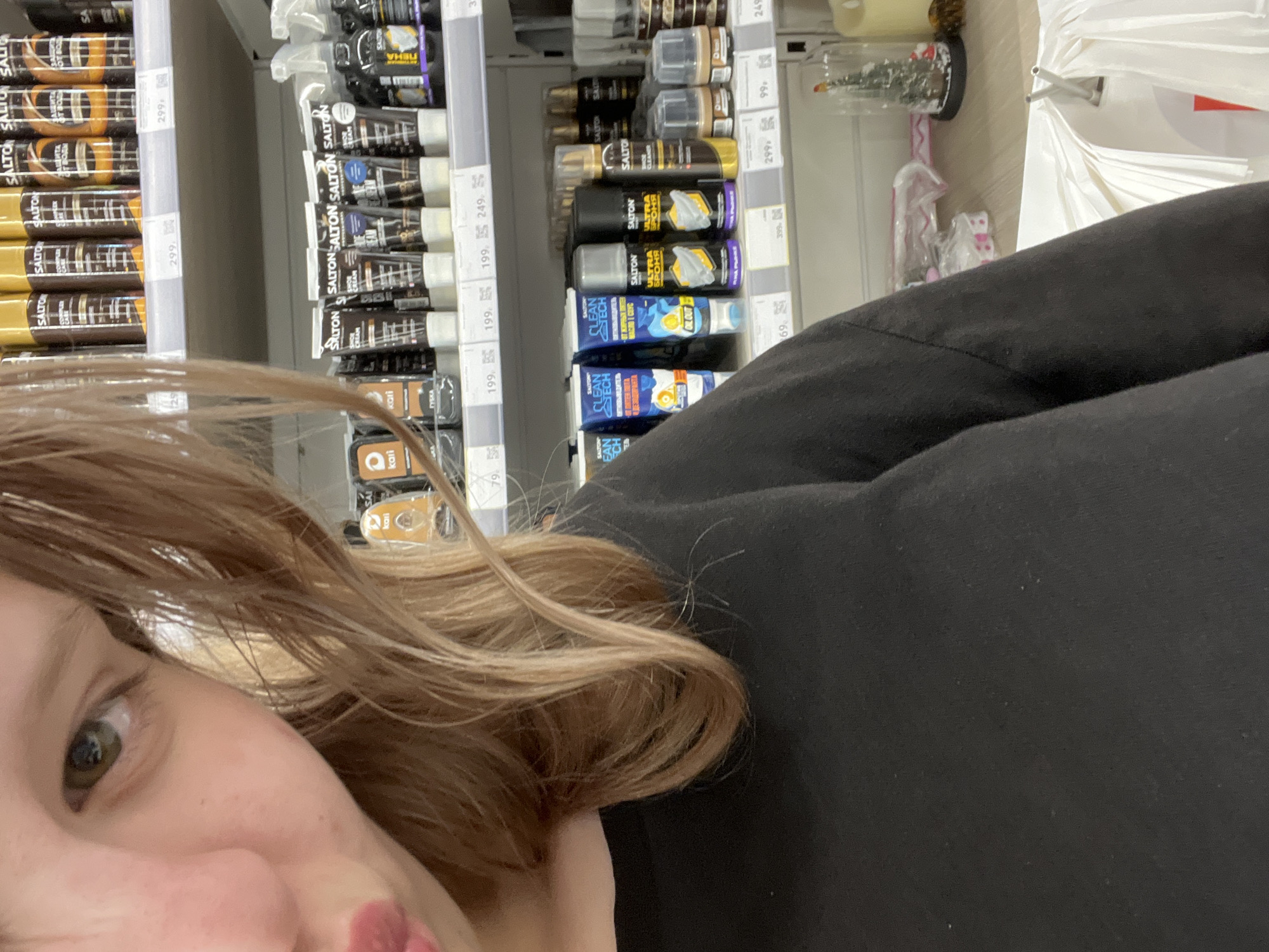 A close-up shot captures the left side of a young woman's face, focusing on her eye and lips, as she looks towards the camera. Her light brown hair is parted on the left and falls across her shoulder. She is wearing a black long-sleeved shirt. The background consists of store shelves displaying various shoe care products, including bottles, tubes, and sprays. Some of the product labels are visible, such as "SALTON" and "CLEAN TECH." Price tags are also present on the shelves. The overall environment appears to be a retail setting, likely a shoe care or cosmetics section of a store. The image's context suggests it was taken in a store located in Kokkorevo, Russia.