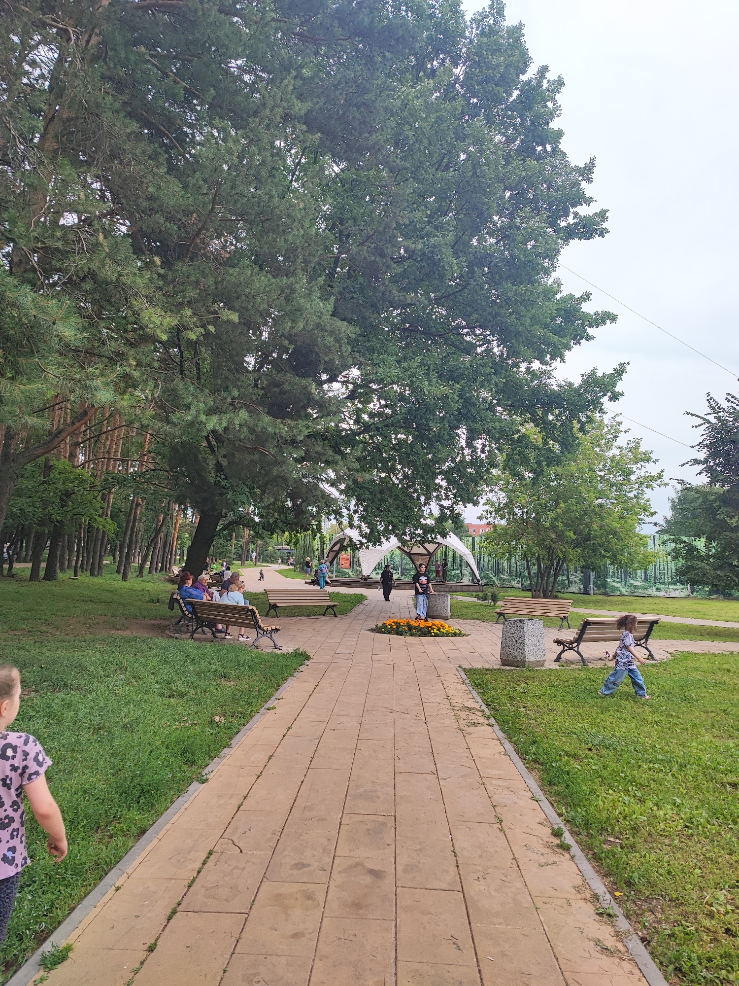 A park scene in Unknown, Unknown shows a wide, paved walkway lined with grassy areas and benches. Several people are present, including a young girl in the foreground walking on the grass and another child running on the grass near a bench. A group of adults are seated on benches on the left side of the walkway. In the background, there is a large tree canopy, a white tent structure, and a patterned backdrop that appears to depict trees. Two trash receptacles are visible along the walkway. The sky is overcast.