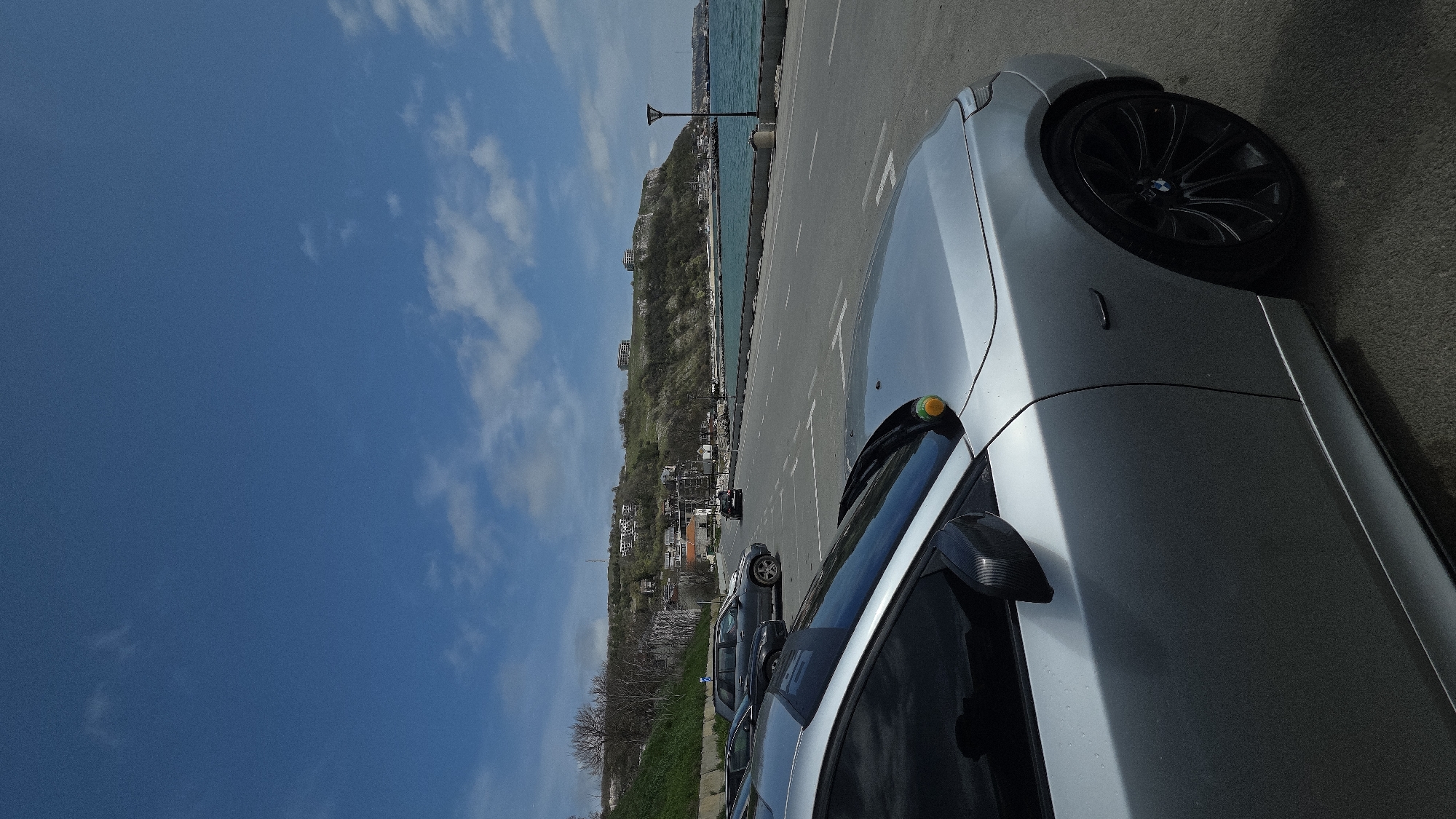 The image depicts a silver BMW parked on the side of a road next to the Black Sea in Kavarna, Bulgaria. The sky is blue with some scattered clouds. In the background, there is a hillside with buildings and trees. Several other cars are parked along the road further back. The foreground focuses on the front quarter of the silver BMW, displaying a black wheel with the BMW logo. The road is paved and marked with white lines.