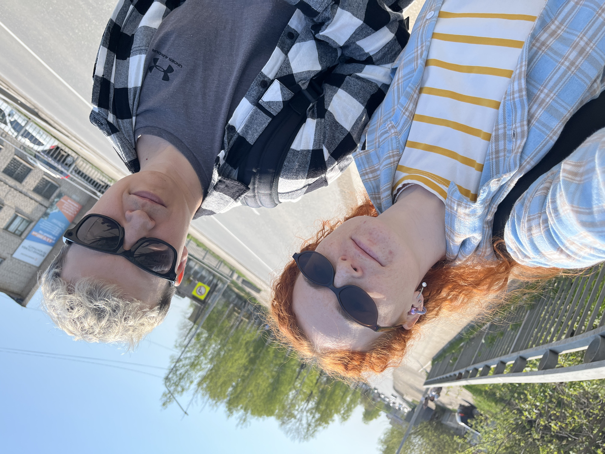Two people are standing outside on a sunny day, close to a bridge with a gray metal railing. The person on the left has short, bleached blonde hair and is wearing black sunglasses, a gray t-shirt with an Under Armour logo, and a black and white checkered flannel shirt. The person on the right has long, curly red hair and is wearing black sunglasses, a white t-shirt with yellow stripes, and a blue and white plaid flannel shirt. In the background, there is a street with cars, buildings, and green trees. There is also a yellow sign with a black arrow on it near the building. The location is Chaikovsky, Russia.