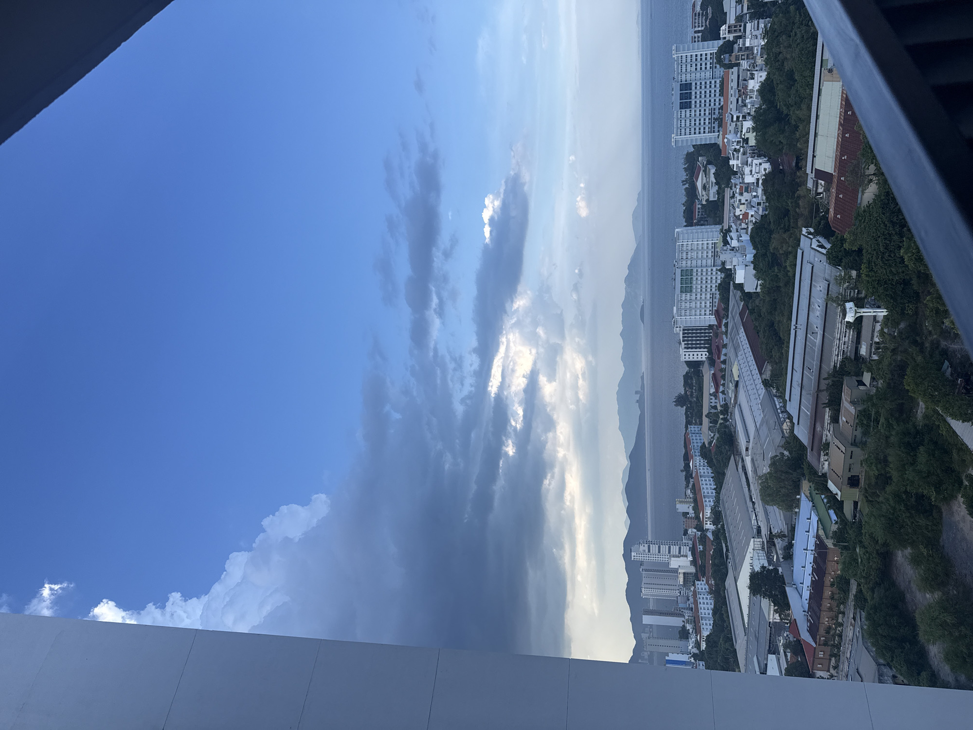 An aerial, wide-angle view of the coastal city of Nha Trang, Vietnam, presents a cityscape dominated by modern architecture and the natural beauty of the sea and mountains. The foreground features the imposing white concrete wall of a building on the left and a dark railing on the right, framing the scene. Beyond these structures, a sprawling urban landscape unfolds, characterized by numerous residential and commercial buildings. High-rise apartment complexes, distinguishable by their white facades and numerous windows, are scattered throughout the city, some standing taller than others. Industrial warehouses with distinctive corrugated metal roofs occupy the mid-ground, interspersed with patches of lush green trees and vegetation.
The city extends to the coast, where the calm, blue waters of the South China Sea meet the land. In the distance, a range of hazy, mountainous islands forms a picturesque backdrop against the horizon. The sky above is a dramatic blend of deep blue and scattered clouds, with patches of bright white cumulus clouds contrasting with darker, more ominous storm clouds gathering on the left, suggesting an approaching weather front. The overall impression is one of a vibrant, developing coastal city situated in a region of significant natural beauty.