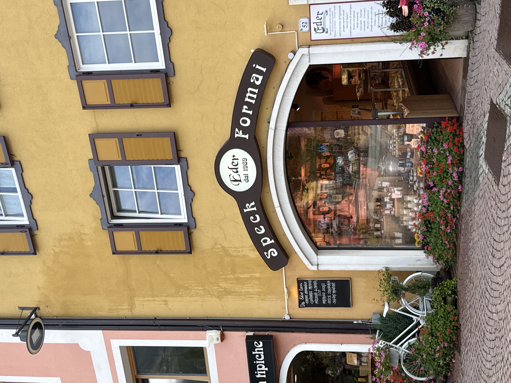 The image shows the exterior of a shop called "Eder Speck & Formai" located in Moena, Italy. The shop is situated on a street with a cobblestone pavement. The building has a yellow facade with brown wooden shutters on the windows. A large, dark brown sign above the shop entrance reads "Eder Speck Formai dal 1989". The shop window displays various food products, likely cheeses and cured meats. In front of the shop, there are flower beds filled with pink and red flowers, and a white bicycle decorated with flowers. To the left of the shop, a pink building with white trim is visible, with a sign that reads "tipiche". To the right of the shop, a vertical sign with the number "52" and the shop's name "Eder" is mounted on the wall. The scene is brightly lit, suggesting daytime. No people are clearly visible in the image.