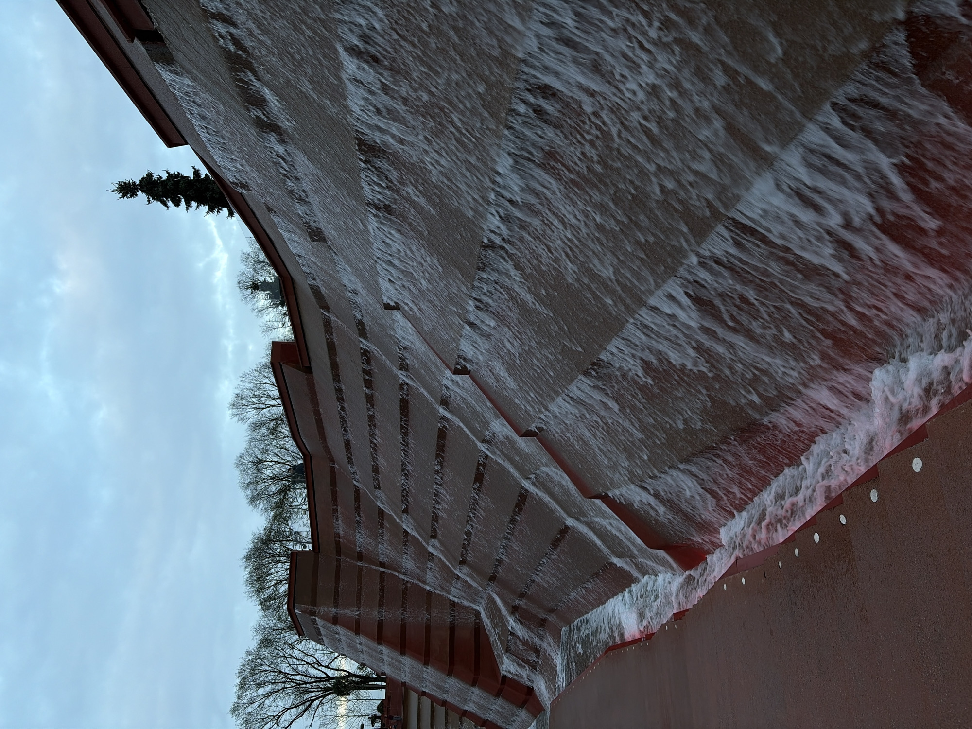The image captures a large, stepped water feature, likely a man-made waterfall, viewed from a low angle looking upwards. A broad, reddish-brown structure forms multiple tiers, down which turbulent water actively cascades. The water is a mix of white foam and clear streams, creating a dynamic texture as it flows over each step. A subtle reddish glow emanates from beneath the water in certain sections, particularly at the lowest visible tier, indicating artificial lighting.
Above the water feature, a light grey, overcast sky dominates the background. Silhouetted against the sky are several trees: a prominent dark evergreen tree (possibly a pine or fir) stands tall on the left, and numerous bare deciduous trees line the horizon on the right, suggesting a late autumn or winter season. Along the top edge of the lowest and largest section of the red structure, a series of evenly spaced circular white or silver objects are visible, which may be decorative elements or recessed lights.
The scene conveys a sense of architectural design combined with natural elements. The time of day appears to be either an overcast day or twilight, emphasized by the artificial light within the water. No individuals or specific identifying text are visible in the image.