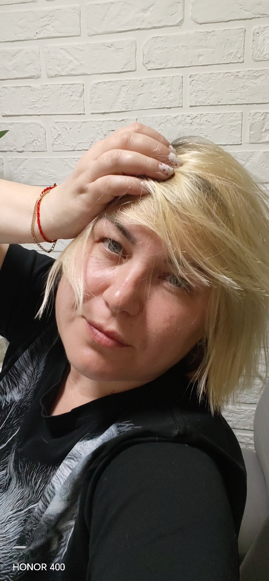 A woman with short blonde hair is posing for a selfie. She is wearing a black t-shirt with a white design. Her left hand is on her head, and she has a red and gold bracelet on her left wrist. The background is a white brick wall. The photo was taken with a HONOR 400 device. There is no explicit indication of the location being Noviy, Russia, in the image.