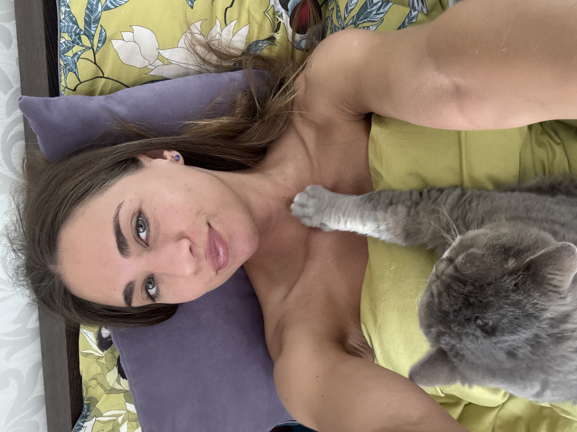 A woman is lying on a bed with a cat. She is on her back, looking at the camera, with her head resting on a purple pillow. She is wearing a yellow top. The cat is lying on her chest, with its paw extended and touching her neck. The bed has a patterned duvet. The scene appears to be indoors, in a bedroom, possibly in Russia.
