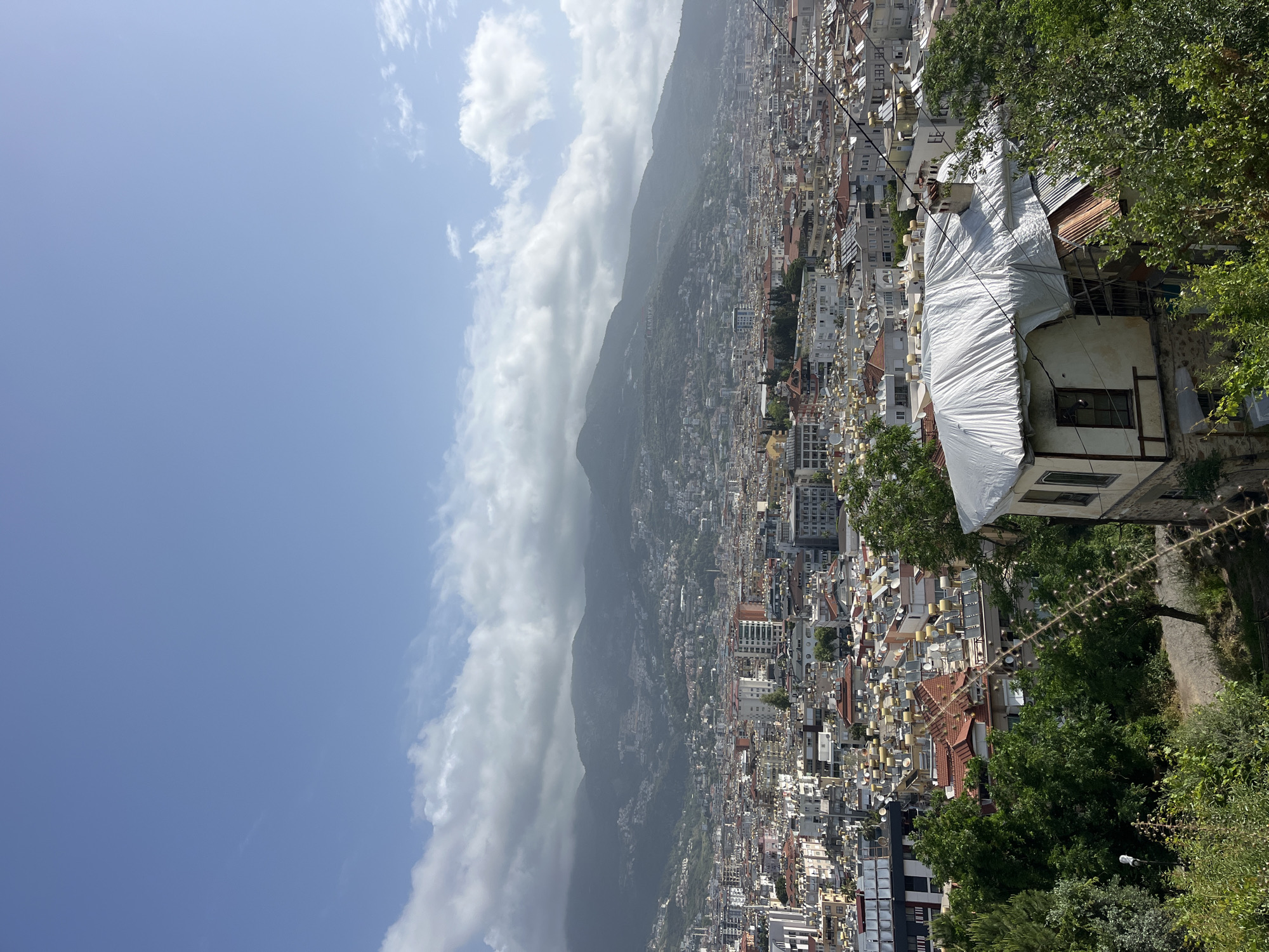 The image depicts a panoramic view of Alanya, Turkey, taken from a high vantage point. In the background, a mountain range is partially obscured by clouds. The foreground features densely packed buildings, with a mix of residential and commercial structures, extending towards the coastline. The buildings are varied in height and color, creating a textured urban landscape. In the right side of the frame, vegetation is visible, suggesting the presence of a park or green space. One notable feature is a building covered in white tarpaulin, possibly undergoing construction or renovation, positioned in the middle right of the image. The sky is blue with scattered clouds. The overall impression is that of a bustling city nestled against a mountainous backdrop.