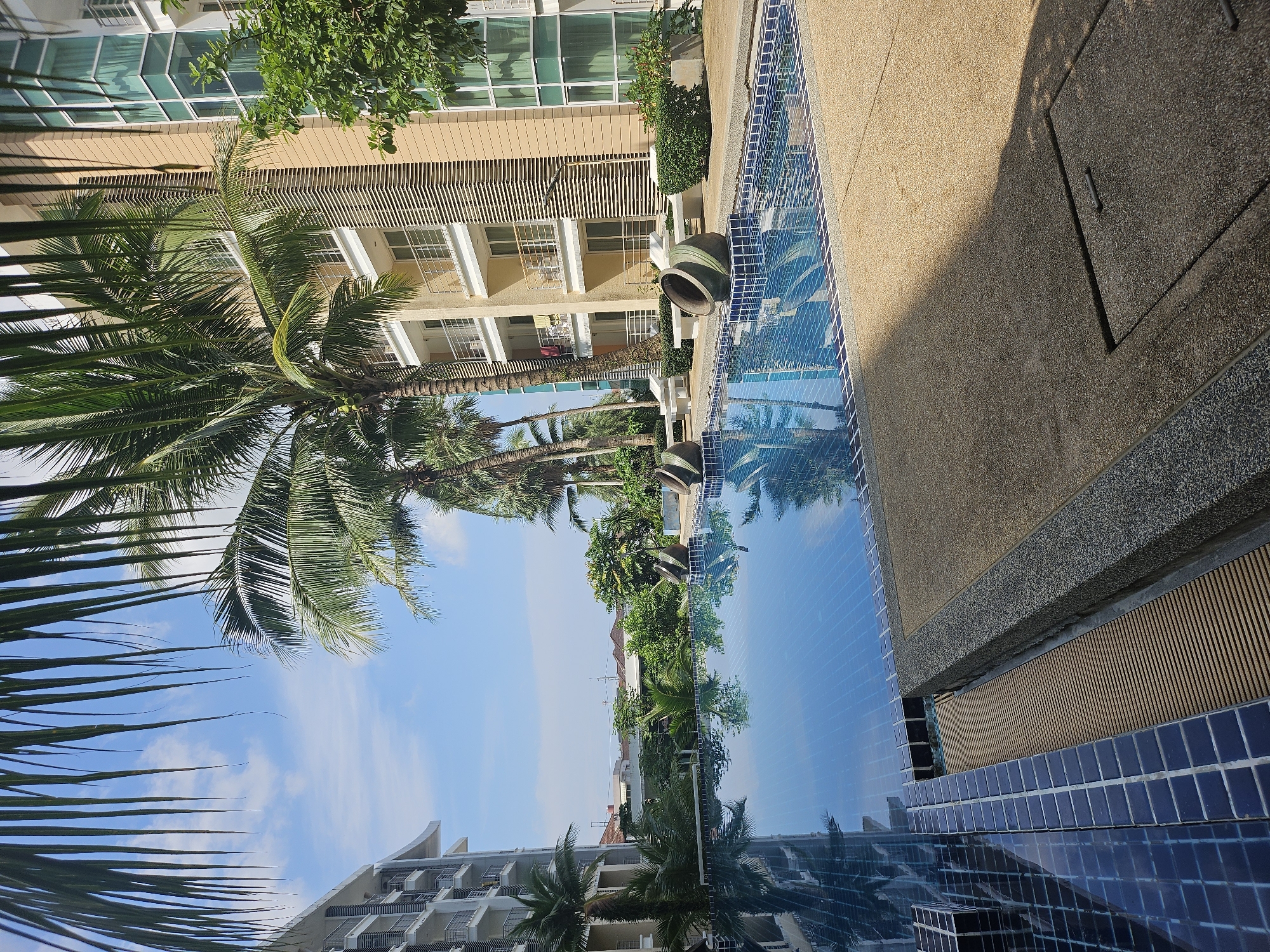 The image depicts a swimming pool area next to a modern apartment building. The pool is rectangular with blue tiled edges and a blue tiled bottom, reflecting the sky and the surrounding environment. Several palm trees are visible, with their fronds partially obscuring the view of the sky and the buildings.
The apartment building is multi-story with cream-colored facade and balconies. A section of the building features a modern glass facade. In the foreground, near the pool, are two large, decorative ceramic pots. The ground around the pool is paved with a sandy-colored, textured material.
The sky is blue with scattered white clouds, indicating a sunny day. The overall scene suggests a resort or residential complex, likely in a tropical or subtropical climate given the presence of palm trees. The image is a static, exterior view of this location. No people are present in the frame. The location appears to be Kathu, Thailand, based on the context provided.