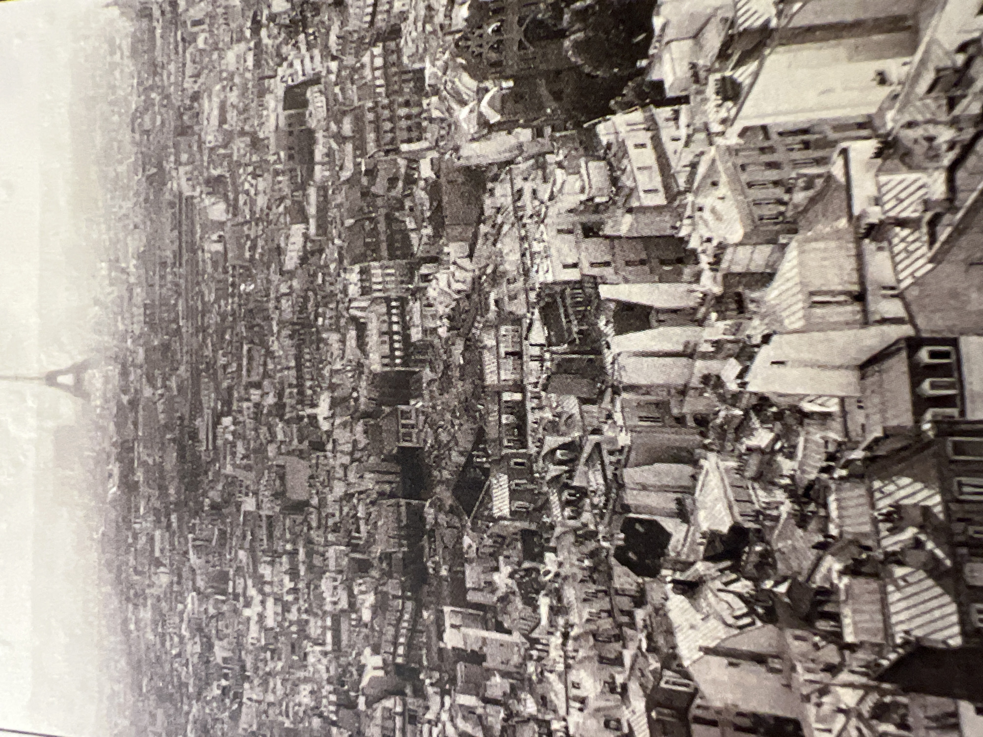 This is a high-angle, black and white photograph of a dense cityscape dominated by buildings with peaked roofs. In the far distance, the Eiffel Tower is visible. The image depicts a sprawling urban environment with numerous tightly packed structures, suggesting a historic city center. The overall tone is hazy, possibly due to atmospheric conditions or the age of the photograph. There are no people or discernible objects in the foreground or midground that allow for specific detail. The absence of any identifying markers or context makes it impossible to pinpoint the location as "Unknown, Russia."