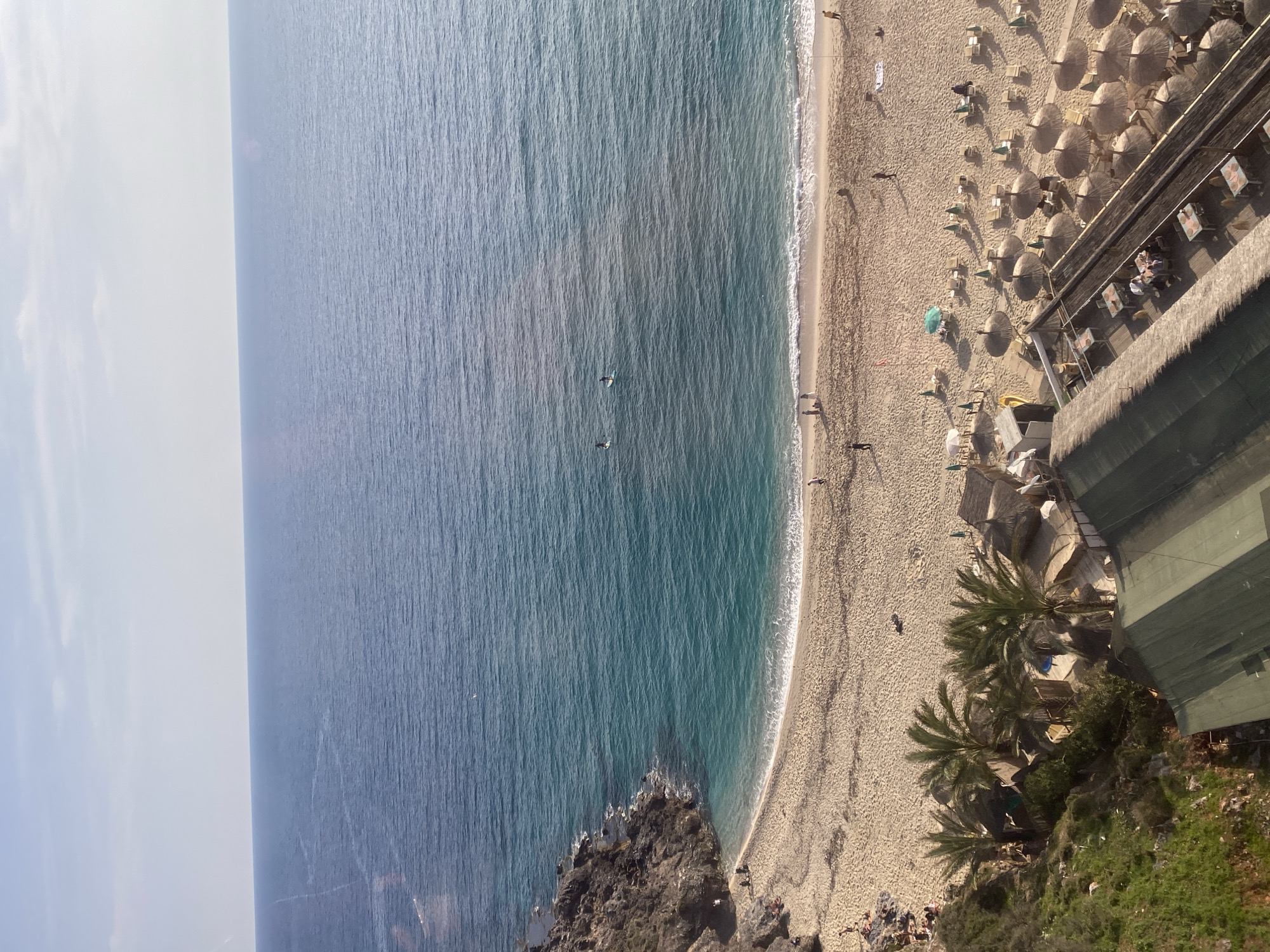 A vertical aerial shot of a beach with a section of the sea next to it. The ocean is dark blue with some light blue areas close to the shore. The beach is sandy with a light brown color. People are scattered on the beach. Several beach umbrellas are arranged in rows. Palm trees are located along the right side of the beach. In the lower left corner, a rocky outcrop extends into the sea. A building is located on the right side of the beach with a green roof. The image is likely taken in Alanya, Turkey.