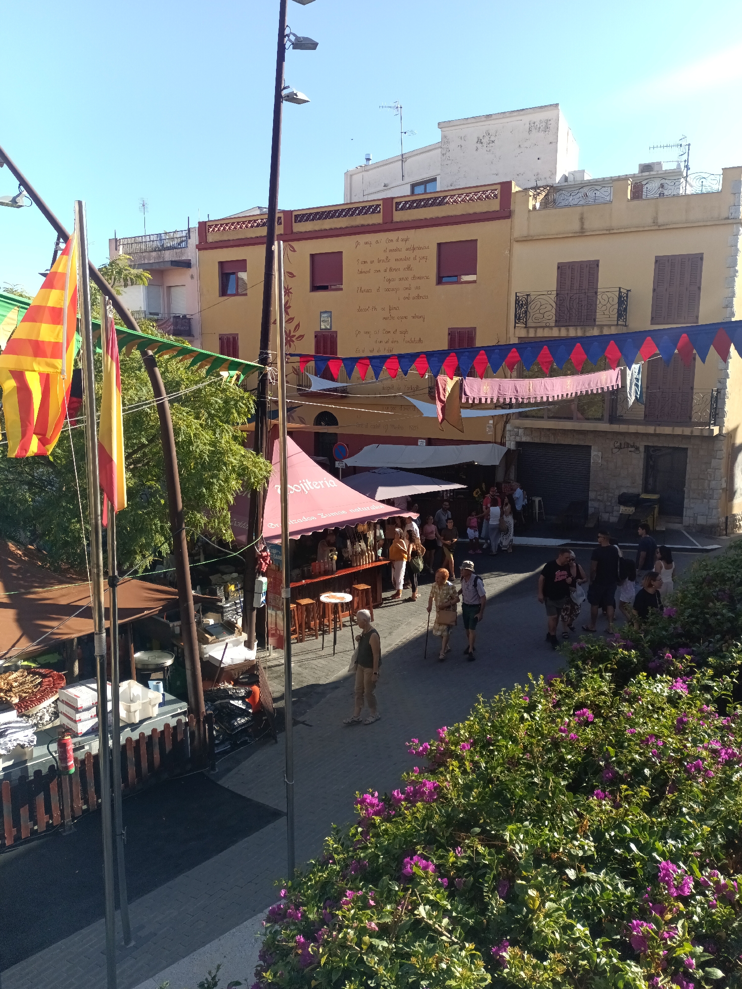 The image depicts a street scene in Calafell, Spain, likely during a local festival or market day. In the foreground, several colorful flags, including the Catalan flag, are prominently displayed, suggesting a celebratory atmosphere. To the left, market stalls are set up, one with a display of food items and another with a red awning that reads "Zumo natural" (natural juice), indicating food and beverage vendors.
People are seen browsing the stalls and walking along the paved street. Some are in casual attire, while others appear to be dressed for the occasion, possibly participating in a historical reenactment or themed event, as suggested by the festive banners and decorations hanging between buildings. The architecture of the buildings shows typical Mediterranean style, with light-colored facades, red-tiled roofs, and balconies. The sky is clear and blue, indicating a sunny day. The presence of lush green bushes with purple flowers in the lower right corner adds a touch of natural beauty to the urban setting. The overall impression is one of a lively community gathering, blending commerce, culture, and leisure in a Spanish town.
