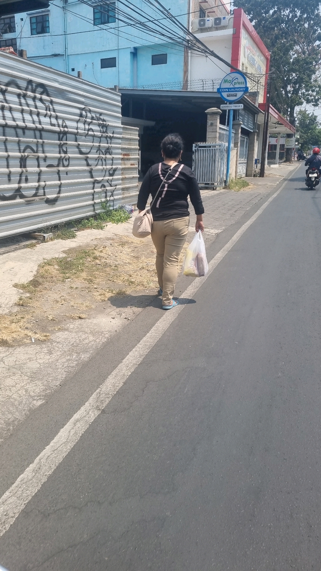 A person is walking along a street in Malang, Indonesia, carrying shopping bags. The person is seen from behind, wearing a black long-sleeved shirt and tan pants, with light blue sandals. They are also carrying a light-colored shoulder bag. The street appears to be paved with asphalt and has a white line marking the edge of the sidewalk. To the left, there is a metal fence with graffiti, and beyond that, a light blue building. To the right, there is a red and white building with a sign that says "Ma Xpress Coin Laundry," indicating a commercial area. In the distance, a motorcycle is visible on the road. The sun is shining, casting shadows on the ground. The overall scene suggests a typical day in a moderately busy urban environment.