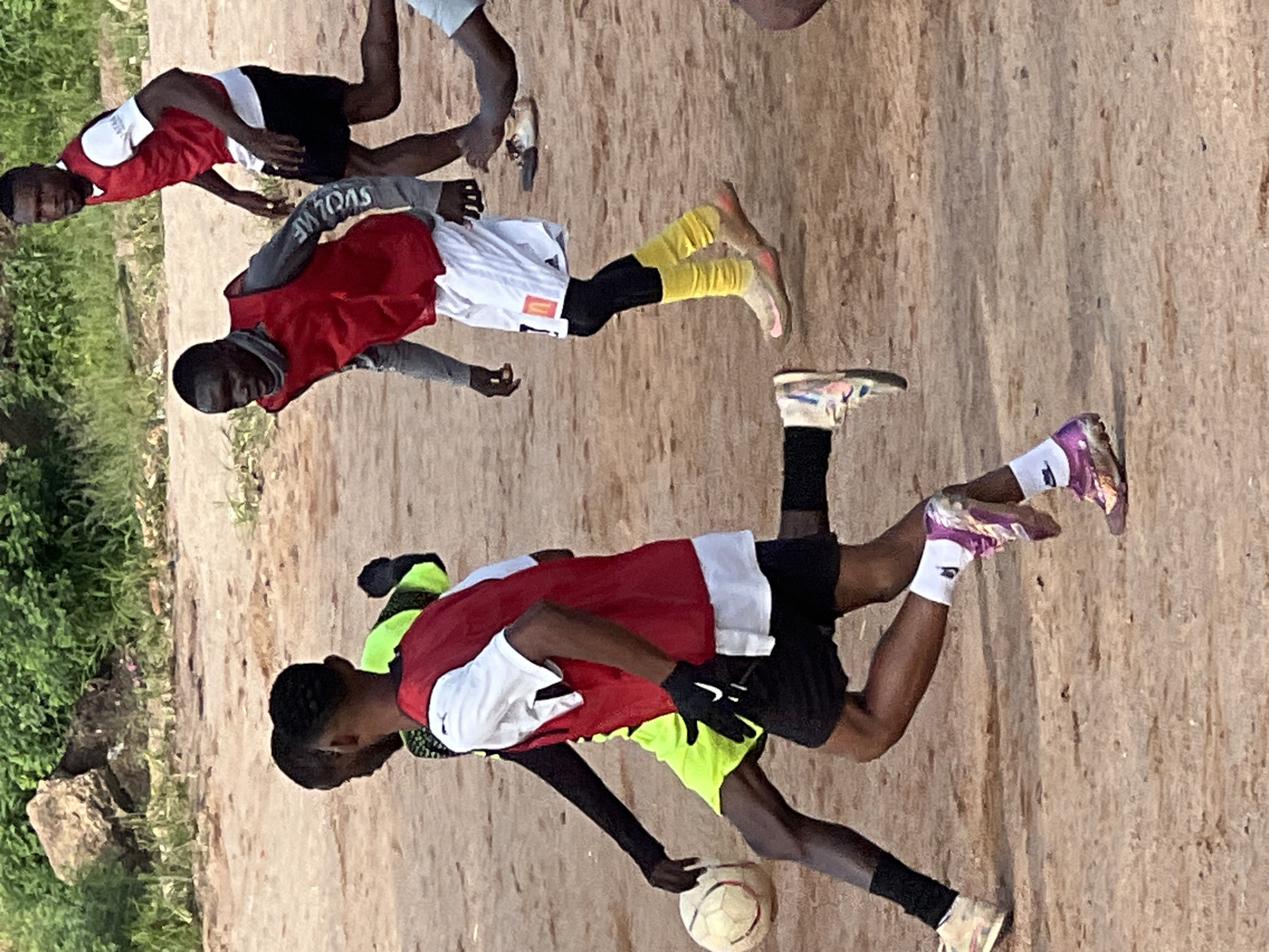 The image shows a group of young men playing soccer on a dirt field, possibly in Jos, Nigeria. The ground is sandy and uneven, with some vegetation in the background. The men are wearing athletic clothing, including vests and shorts. One man is wearing a bright yellow shirt and black shorts, while another is wearing a red vest over a white shirt and black shorts. A third player is wearing a red vest over a grey long-sleeved shirt and white shorts. They are actively engaged in the game, with one player kicking the ball, another dribbling, and others in pursuit. The lighting suggests it is daytime.