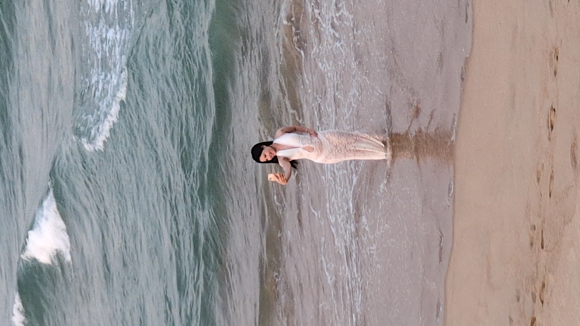 A woman stands in the shallow water at the edge of a beach. She is wearing a light-colored, long dress. She is holding a mobile phone, possibly taking a picture. The water is washing around her legs and the lower part of her dress. The beach is composed of light-colored sand. The ocean extends into the background, with waves visible. The location is likely in Tuy Hòa, Vietnam, based on context.
