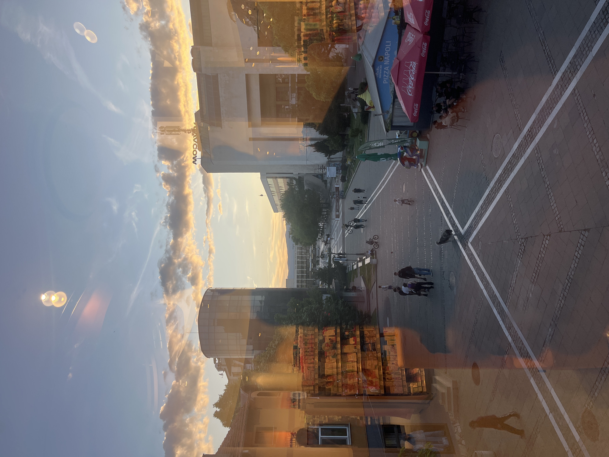 The image shows a cityscape of Blagoevgrad, Bulgaria, likely viewed from an elevated position through a window. The scene is partially obscured by reflections on the glass. The street-level view features a pedestrian area with geometric pavement patterns defined by white lines. People are seen walking and seated at outdoor tables under Coca-Cola and Pizza Napoli umbrellas. A large building with the word "AVICOW" on its side is visible, along with a cylindrical building made of glass. The sky is visible with clouds. The reflections on the glass show a building with many orange colored boxes, and other street-level views in Blagoevgrad. The lighting indicates it is either sunrise or sunset.