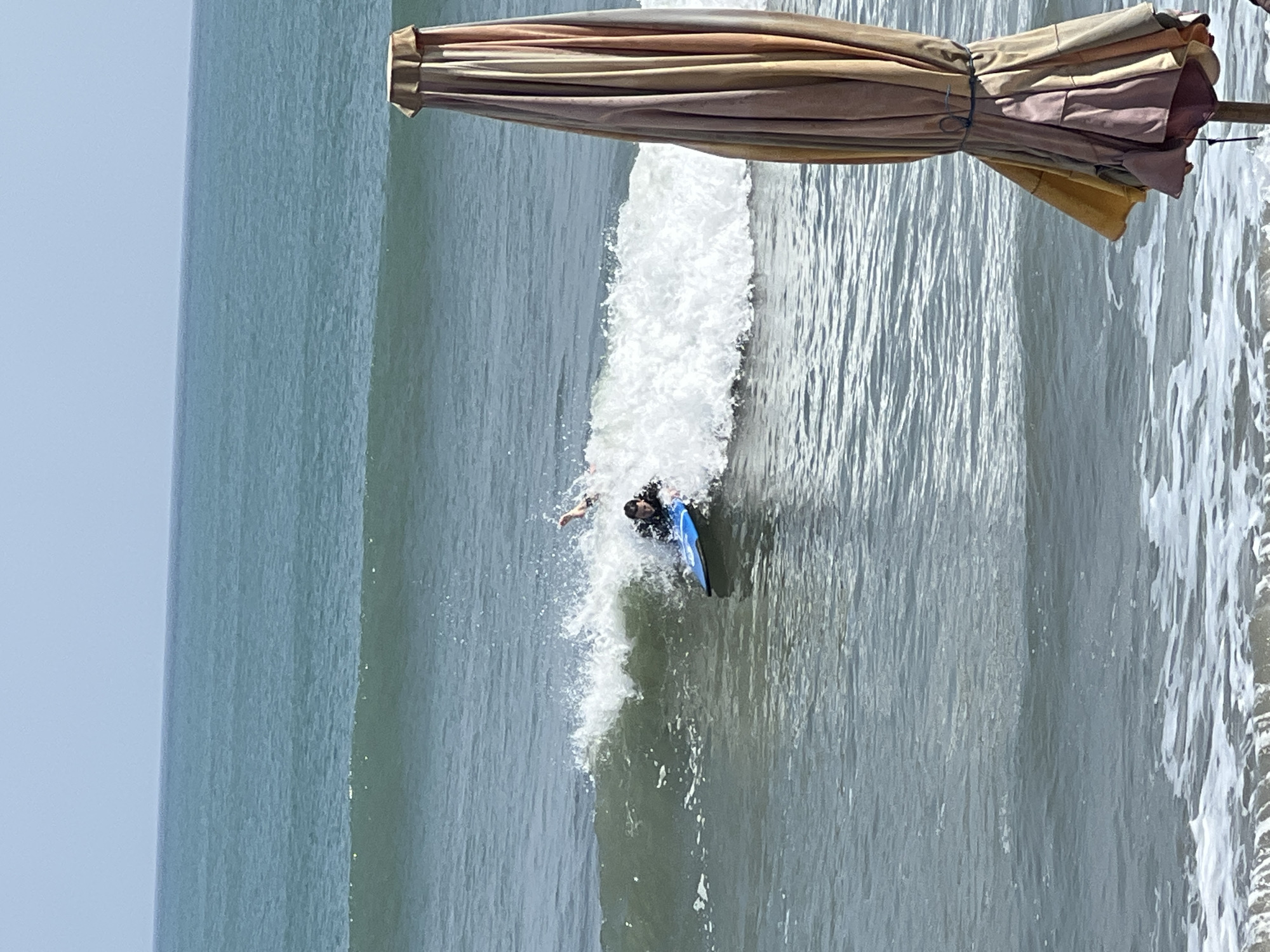 A person is bodyboarding in the ocean in Kuta, Indonesia, amidst crashing waves and white foam. The person is wearing a black rash guard and is positioned on a blue bodyboard, actively riding a wave. Another person's legs can be seen in the water further behind, suggesting they are also participating in water sports. In the foreground, a partially closed beach umbrella with striped fabric stands on the right side of the frame, adding a detail of beach amenities to the scene. The ocean water is a teal color, with ripples and waves indicating moderate sea conditions. The sky above is clear and a pale blue.