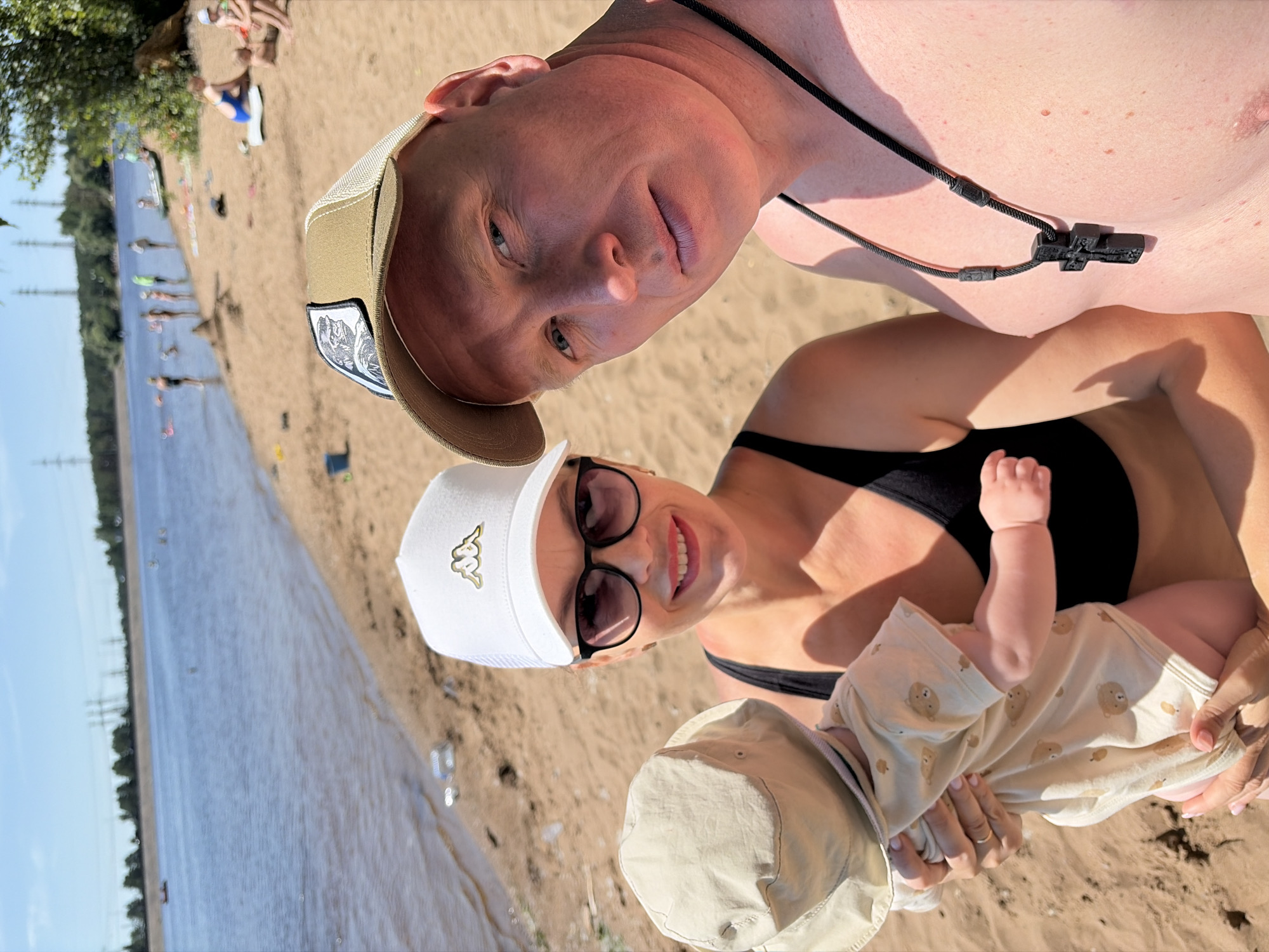In Chaikovsky, Russia, a family is enjoying a sunny day at the beach. A man and a woman are posing for a selfie, with the woman holding a baby. The woman is wearing a white Kappa hat and sunglasses, while the man is wearing a tan trucker hat and a cross necklace. The baby is wearing a sun hat and a onesie with a bear print. The background shows a body of water, a sandy beach, and several other people enjoying the day. The overall scene is one of leisure and family fun.