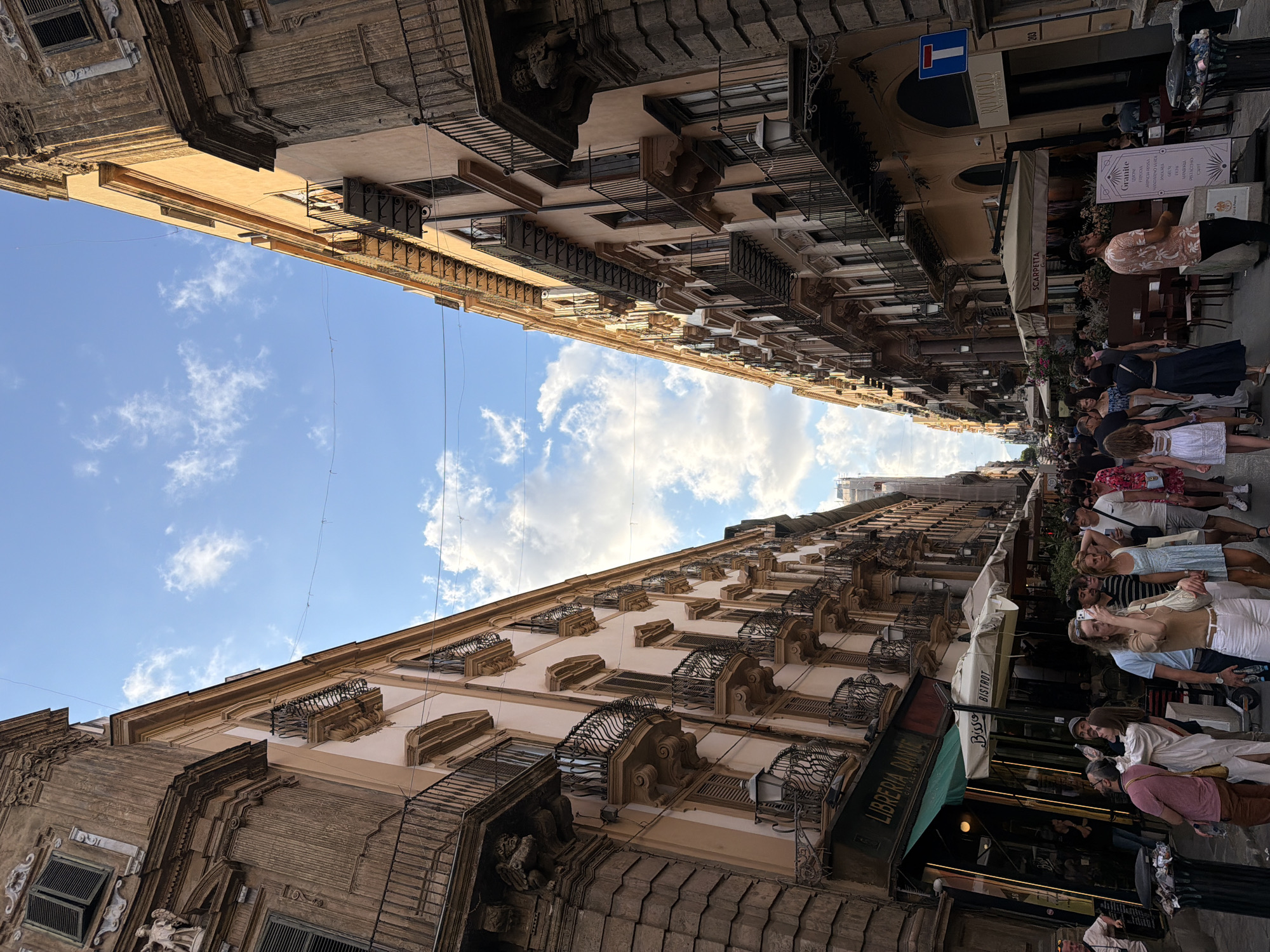 The image shows a bustling street scene in Palermo, Italy. The perspective is looking down a narrow street lined with tall, ornate buildings on either side. The architecture features balconies with wrought-iron railings and classical decorative elements. The sky above is blue with scattered white clouds.
In the foreground, people are walking along the street and patronizing outdoor seating areas of businesses. Several cafes and shops are visible, with awnings and signage indicating their presence. One shop has a sign that reads "LIBRERIA DANTE." People are dressed casually, suggesting warm weather. Some individuals are taking photos with their phones, while others are engaged in conversation or walking.
The overall impression is a lively urban environment, characteristic of a European city with historical architecture. The lighting suggests it is daytime, likely late afternoon given the angle of the sun casting shadows.
