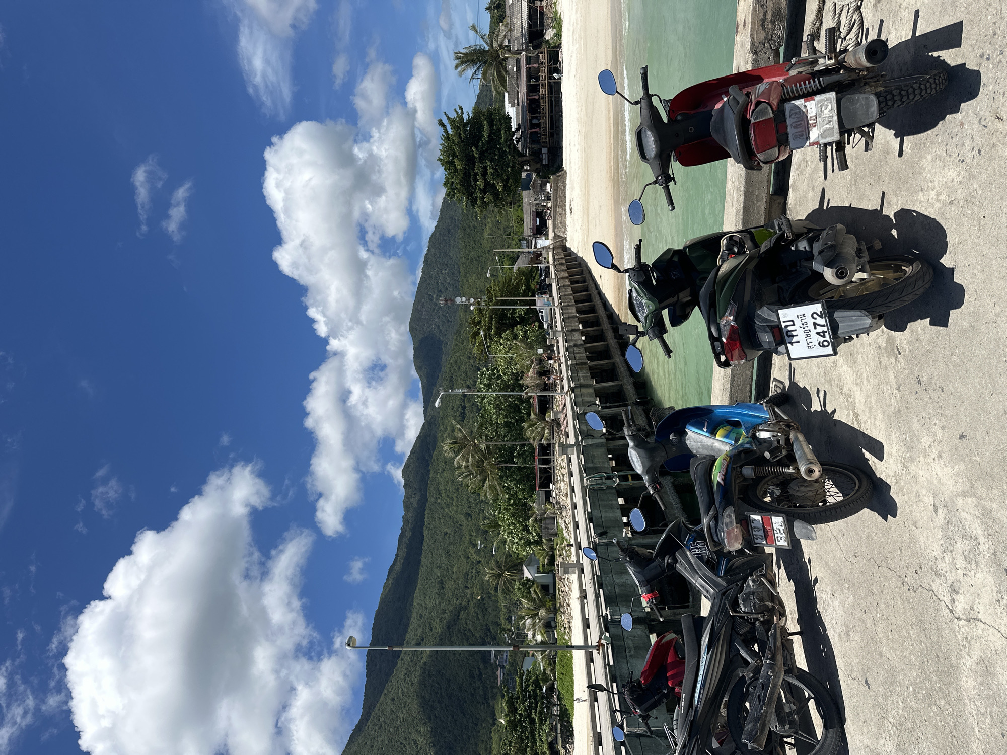 In ตำบลเกาะพะงัน, Thailand, a row of five motorcycles is parked on a concrete pier. The motorcycles, facing away from the camera, are positioned along the edge of the pier. The pier extends over a body of turquoise water. In the background, a sandy beach lines the shore, backed by lush green hills and mountains under a bright blue sky dotted with white clouds. A wooden pier structure with railings and lampposts is visible behind the parked motorcycles. Beyond the beach, a few buildings with thatched roofs suggest a coastal settlement.