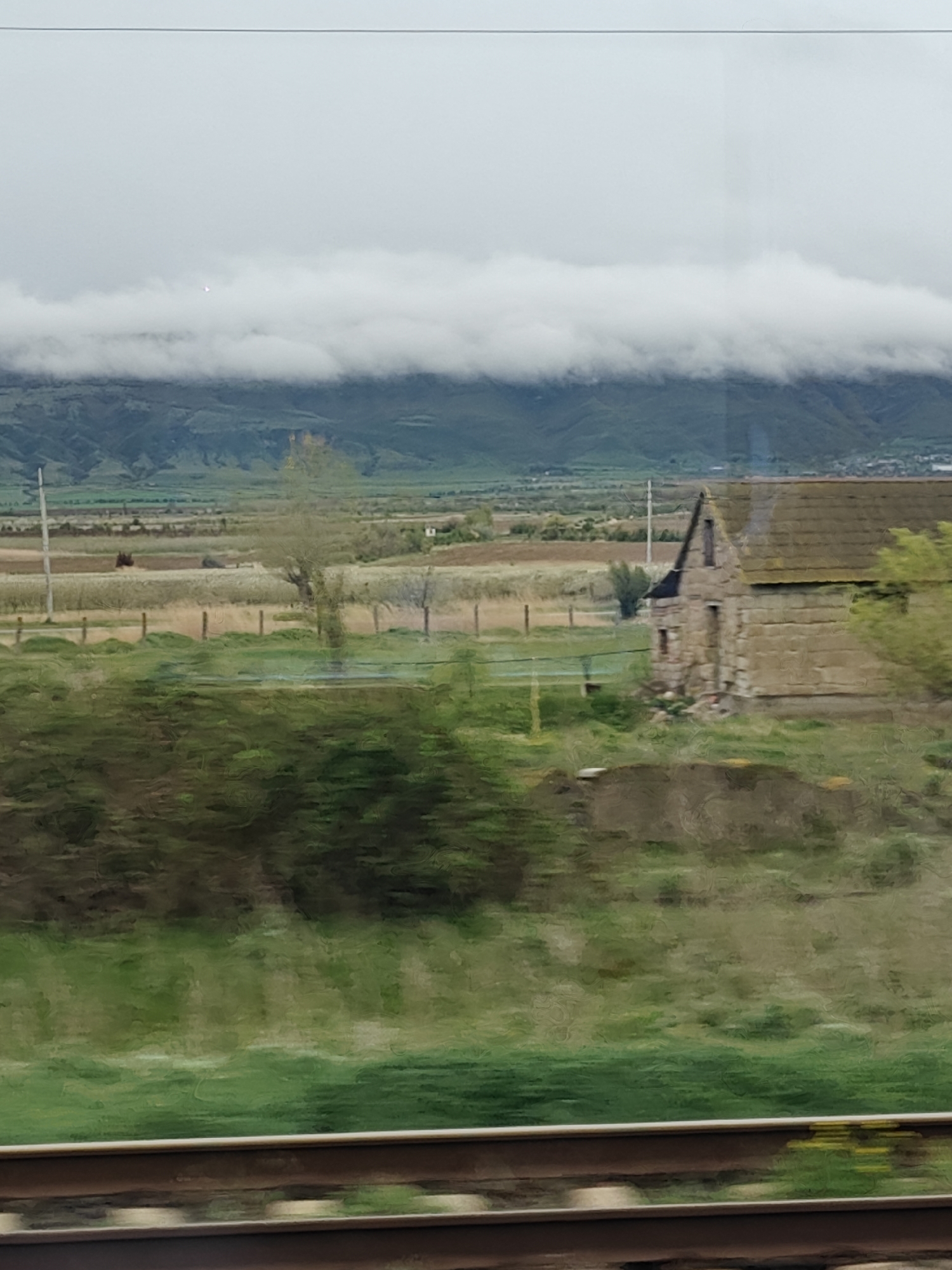 The image depicts a landscape scene viewed from a moving train, indicated by the blurred foreground and visible railway tracks. A dilapidated stone structure with a gabled roof stands to the right of the frame, near a small, bare tree. Beyond this structure is a field enclosed by a simple fence with wooden posts. In the background, rolling hills are visible, shrouded in low-lying clouds or fog. A few power lines are seen extending across the open landscape. The sky is overcast. The location is not specified, and no people or specific interactions are evident in the image. The environment is rural or agricultural, with evidence of human modification.