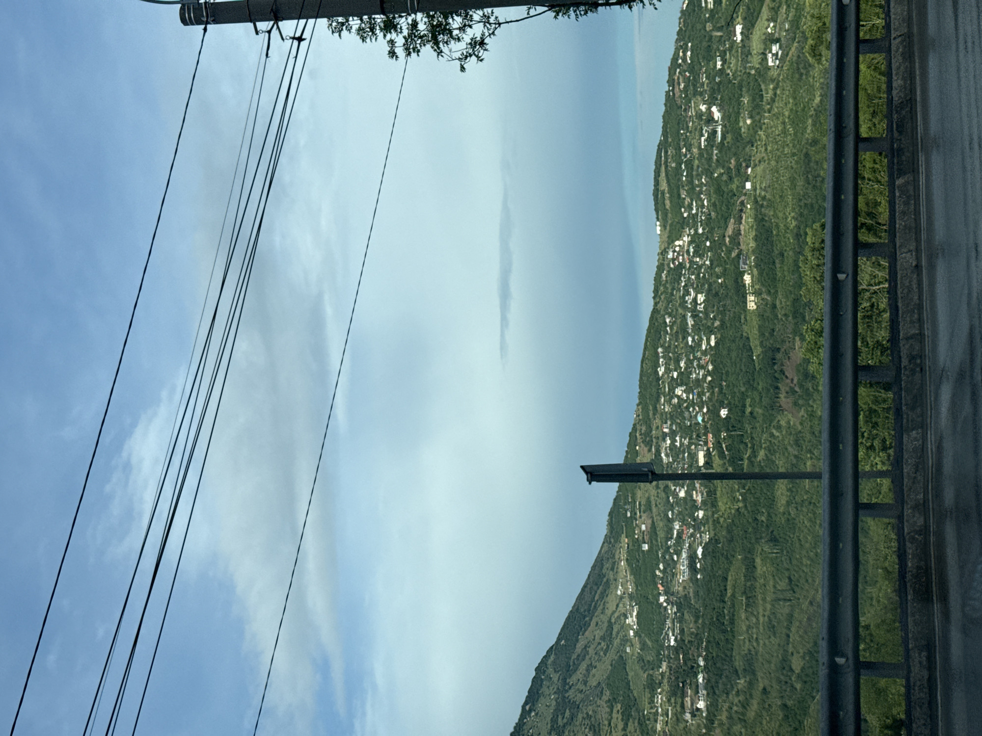 The image shows a view of a hillside residential area. The hillside is covered in green vegetation and dotted with numerous houses. In the foreground, there is a road with a metal guardrail. Several power lines are visible strung across the sky. A utility pole is also present on the right side of the image. A signpost stands near the center of the frame. The sky is partly cloudy and blue.