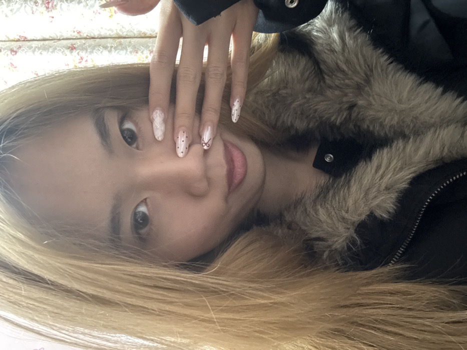 A female subject is depicted in a close-up indoor photograph, located in Shapingba District, China. The subject, with long light-brown hair, looks directly forward with a slight smile. She wears a dark jacket featuring a prominent light-brown faux fur collar. Her left hand is raised, partially obscuring the lower right quadrant of her face. Her fingernails are manicured; the index finger displays a white textured design, the middle and pinky fingers show dark polka dots on a light base, and the ring finger features a red bow design. The background consists of a light-colored curtain with a subtle floral pattern.