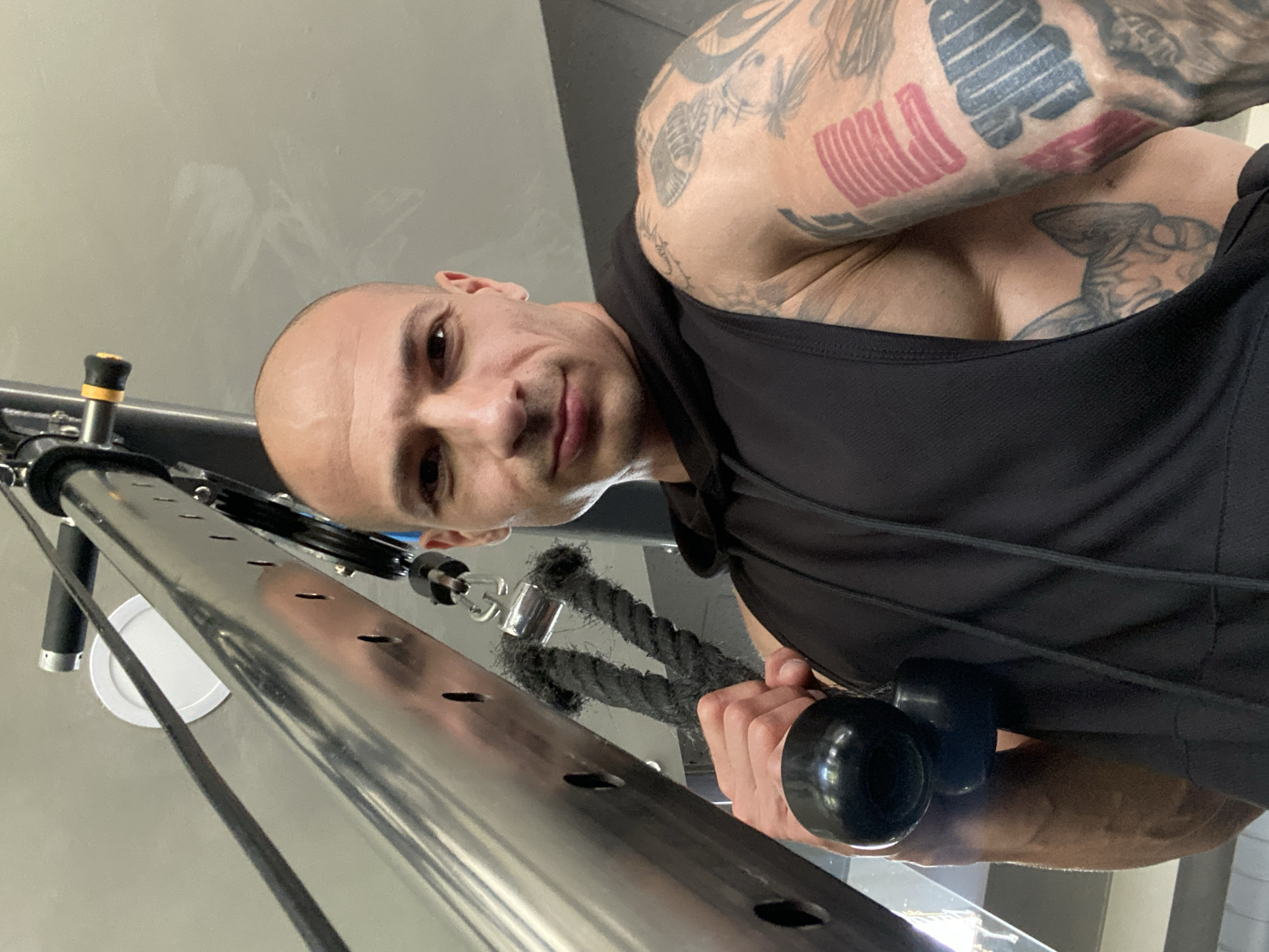 A man is holding a cable attachment on a gym machine. The man has a shaved head and tattoos on his arms. He is wearing a black tank top with a hood. The gym equipment is silver and black. The background is a light gray wall. The location is not specified in the image.