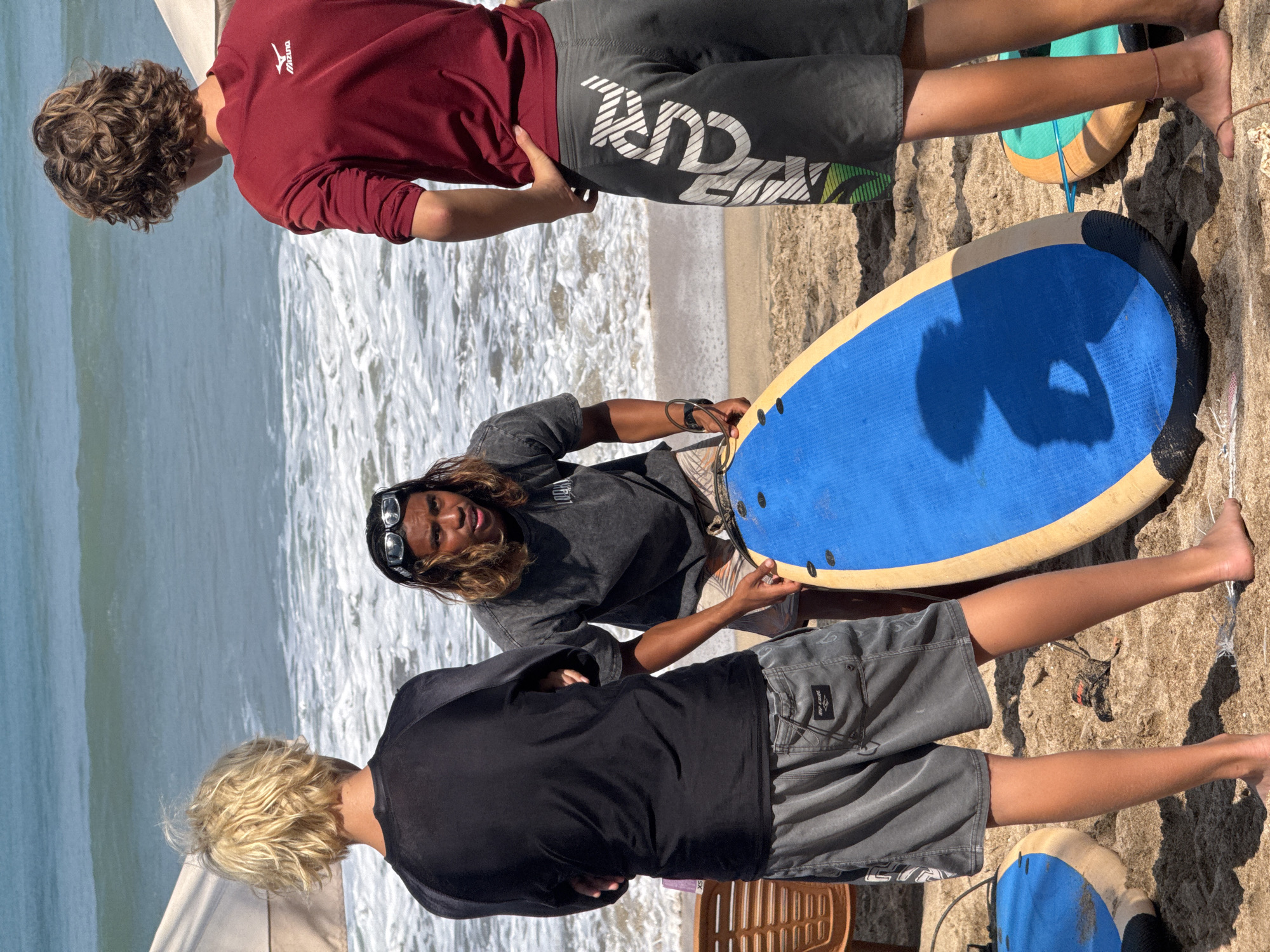 Three individuals, likely young men, are on a sandy beach in Kuta, Indonesia, with the ocean and waves visible in the background. Two are facing away from the camera, one in a black rash guard and grey shorts, and the other in a red long-sleeved shirt and grey shorts with white lettering. The third individual, facing the camera, is wearing a dark t-shirt and patterned swim trunks. They are holding a blue and yellow surfboard. The context suggests a surfing lesson or outing.