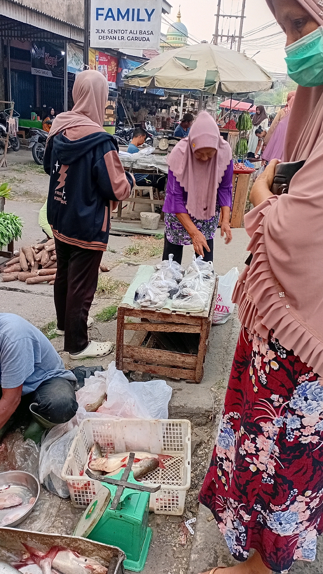 The image depicts a bustling street market scene in Jambi, Indonesia, likely in the vicinity of Jalan Sentot Ali Bassa, as indicated by a sign for "FAMILY" with this address and "DEPAN LR. GARUDA" (in front of Garuda Lane). The foreground is dominated by a display of fresh fish laid out on a green mechanical scale and in plastic crates, suggesting a food market. Several individuals are engaged in market activities: a person in a blue shirt is partially visible and appears to be working near the fish, while another individual wearing a pink hijab and purple top is attending to a wooden crate filled with bagged items, possibly more fish or other goods for sale. Another person in a dark jacket and light pink hijab stands nearby, looking towards the market stalls. In the background, more market stalls are visible under canopies, with umbrellas providing shade. The overall atmosphere is that of a typical Indonesian wet market, with people engaged in commerce amidst various goods and produce. The presence of motorbikes parked on the side indicates it is an urban setting.