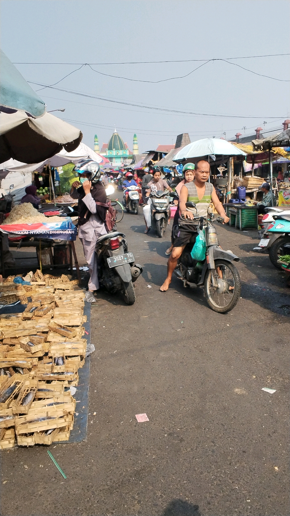This is a full, eye-level, outdoor, daytime photograph of a bustling street market in Mojokerto, Indonesia. The scene is crowded with people, motorcycles, and market stalls. In the background, a large mosque with a green dome and two minarets is visible. The foreground is dominated by a pile of wooden crates filled with dried fish.
In the center of the image, a man with a shaved head, wearing a grey and white striped sleeveless shirt and black shorts, is riding a dark-colored motorcycle. He is barefoot. Behind him, a woman wearing a green helmet and a light-colored shirt is also on the motorcycle. To the left of this motorcycle, a woman in a grey hijab and grey pants stands next to a dark-colored motorcycle with a license plate that reads "S 3527 TG 10-21". She is wearing a dark helmet and appears to be adjusting something on her face. In front of her, a market stall is laden with piles of what appears to be dried fish in wooden crates.
Further back in the scene, other individuals on motorcycles are navigating the street. There are market stalls with various goods, and umbrellas providing shade. The road surface is asphalt. The sky is clear and pale blue. The overall atmosphere suggests a busy, active marketplace.