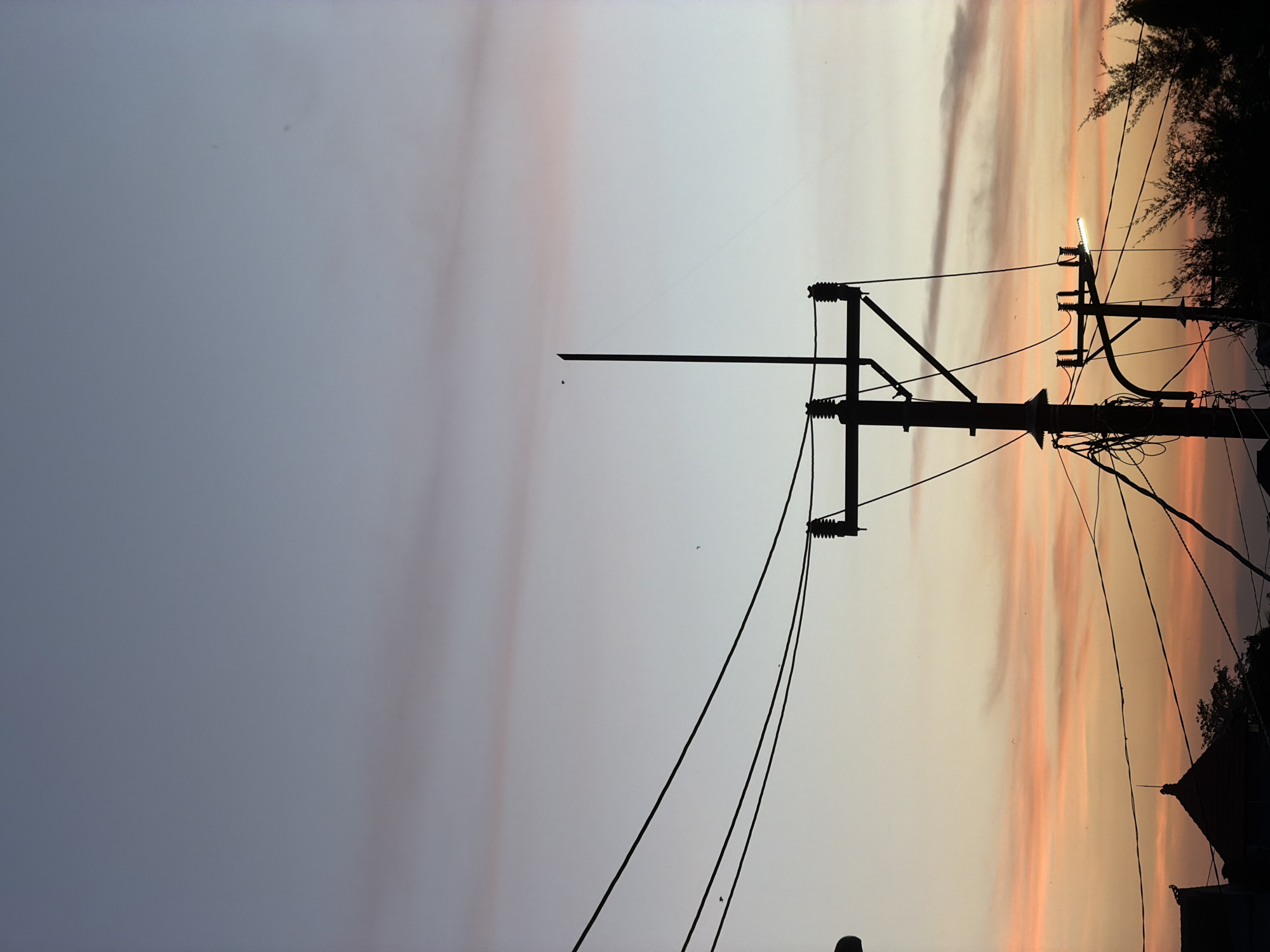 The image depicts a silhouette of electrical poles and wires against a sunset sky in North Kuta, Indonesia. The sky exhibits a gradient of colors from light greyish-blue to pale orange with pink streaks. The electrical poles are the prominent feature, with multiple wires extending in various directions. Some foliage and the silhouette of a building can be seen on the right side of the image.
