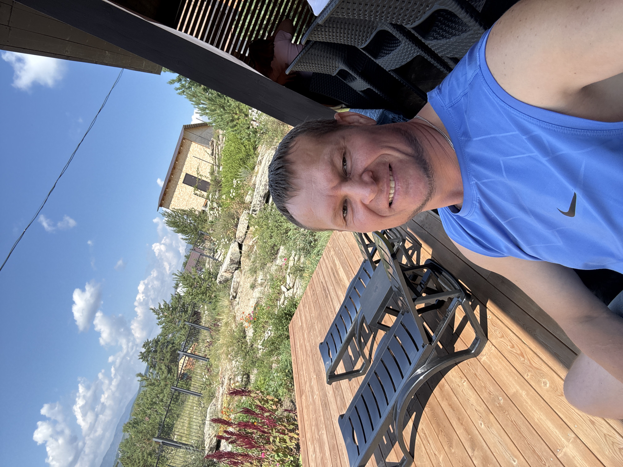 The image is a selfie taken outdoors on a sunny day. The foreground is dominated by a wooden deck with two black lounge chairs. A man, likely in his 40s, is seated and looking at the camera, smiling. He is wearing a blue sleeveless shirt. In the background, there is a natural landscape with green trees, rocks, and a partially constructed building. A black fence runs along the edge of the property. The sky is blue with scattered white clouds. The location is stated to be in New Bukhtarma, Kazakhstan.