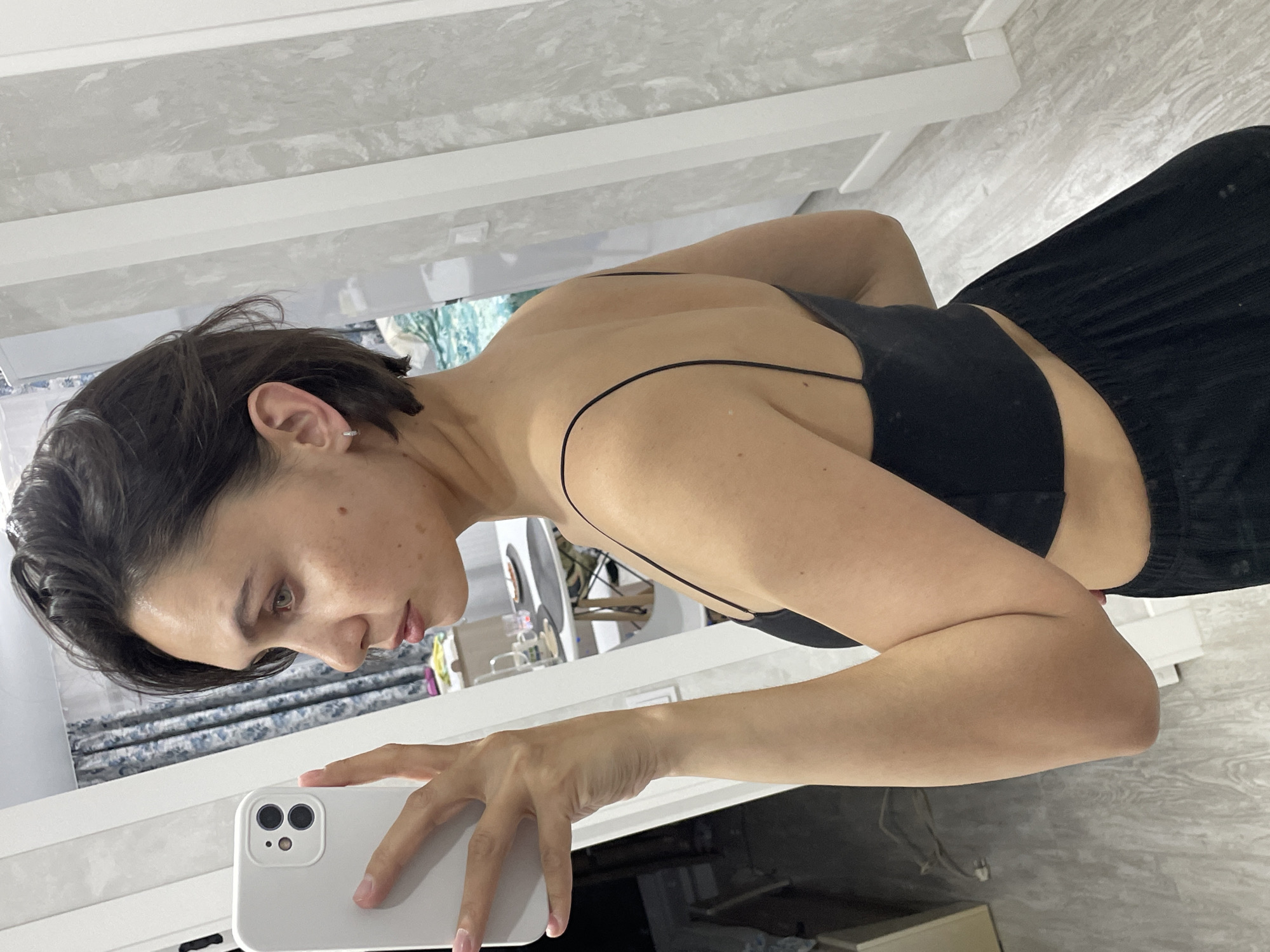 A woman with short, dark hair is taking a selfie in a bright, indoor environment. She is wearing a black top with thin straps and a black skirt or pants. Her left arm is extended, holding a white smartphone with a dual-lens camera. The room appears to be a bathroom or vanity area, featuring white countertops and a decorative mirror reflecting the scene. The location of the scene is unknown; there is no evidence to indicate Izhevsk, Russia.