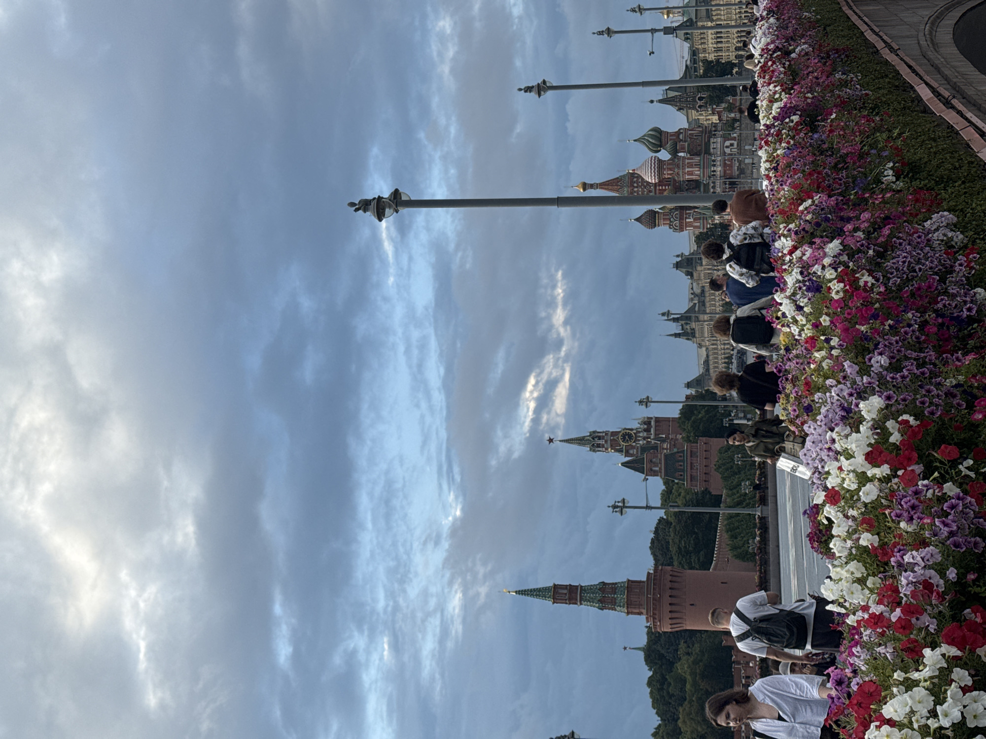 The image captures a view of the Moscow Kremlin and Saint Basil's Cathedral under a cloudy sky, as seen from a bridge adorned with a profusion of colorful flowers. In the foreground, a diverse group of individuals are walking across the bridge, some facing away from the camera, others in profile. They are dressed in casual attire, with some carrying backpacks. The flower beds create a vibrant border along the edge of the bridge. In the background, the distinct red brick towers of the Kremlin, including Spasskaya Tower with its clock, are visible, along with the iconic onion domes of Saint Basil's Cathedral. The sky is filled with dramatic, layered clouds, suggesting either the early morning or late afternoon light. The overall scene depicts a bustling urban environment with historical landmarks set against a natural, floral display.