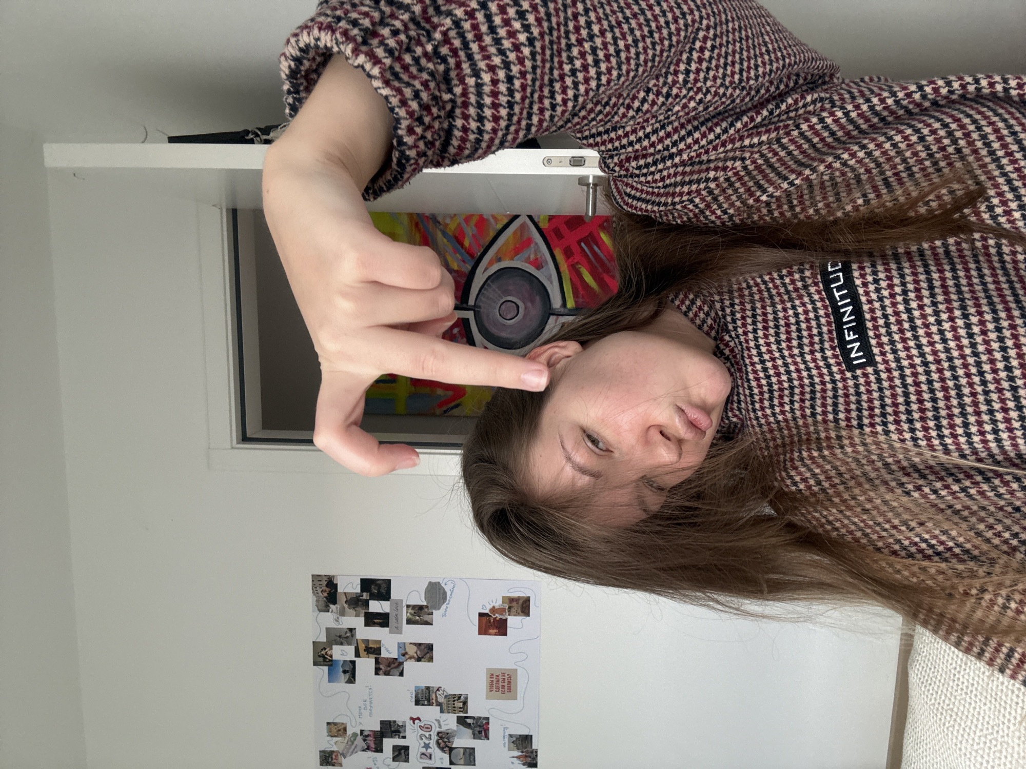 The image depicts a young female in an indoor residential setting. The subject, positioned centrally, is a female with long, light brown hair, estimated to be in her late teens or early twenties. She is wearing a high-necked top featuring a black, brown, and beige houndstooth pattern, with a black rectangular patch on her left chest displaying the white capitalized text "INFINITUD." Her head is tilted to her right, and her lips are pursed in a slight pout. Her right hand is raised, with fingers bent, and the index finger points downwards towards her right ear, partially obscuring her face.
The background consists of a plain white wall. To the left, a white board is affixed, covered with numerous small photographs, some arranged in a grid, and handwritten notes. Visible English text includes "2+2=6" and "I wish love." Cyrillic text is also present, including phrases such as "У МЕНЯ ВСЕ ПОЛУЧАЕТСЯ!", "Я ВСЁ ПРОБОВАЛА!", and "чтобы вы охуели если вы не боитесь!". To the right, a white door frame reveals a brightly colored, abstract painting in an adjacent room, featuring a large stylized eye with a dark pupil and iris, against a background of yellow, red, and blue brushstrokes. The door itself has a silver-colored handle and a locking mechanism. In the lower left foreground, a segment of a light-colored, textured fabric, possibly a blanket or bedspread, is visible.
Based on the visual evidence, the specific location within the city of Vozdovac Urban Municipality, Serbia, cannot be determined from this image. The scene is consistent with a private residential interior.
