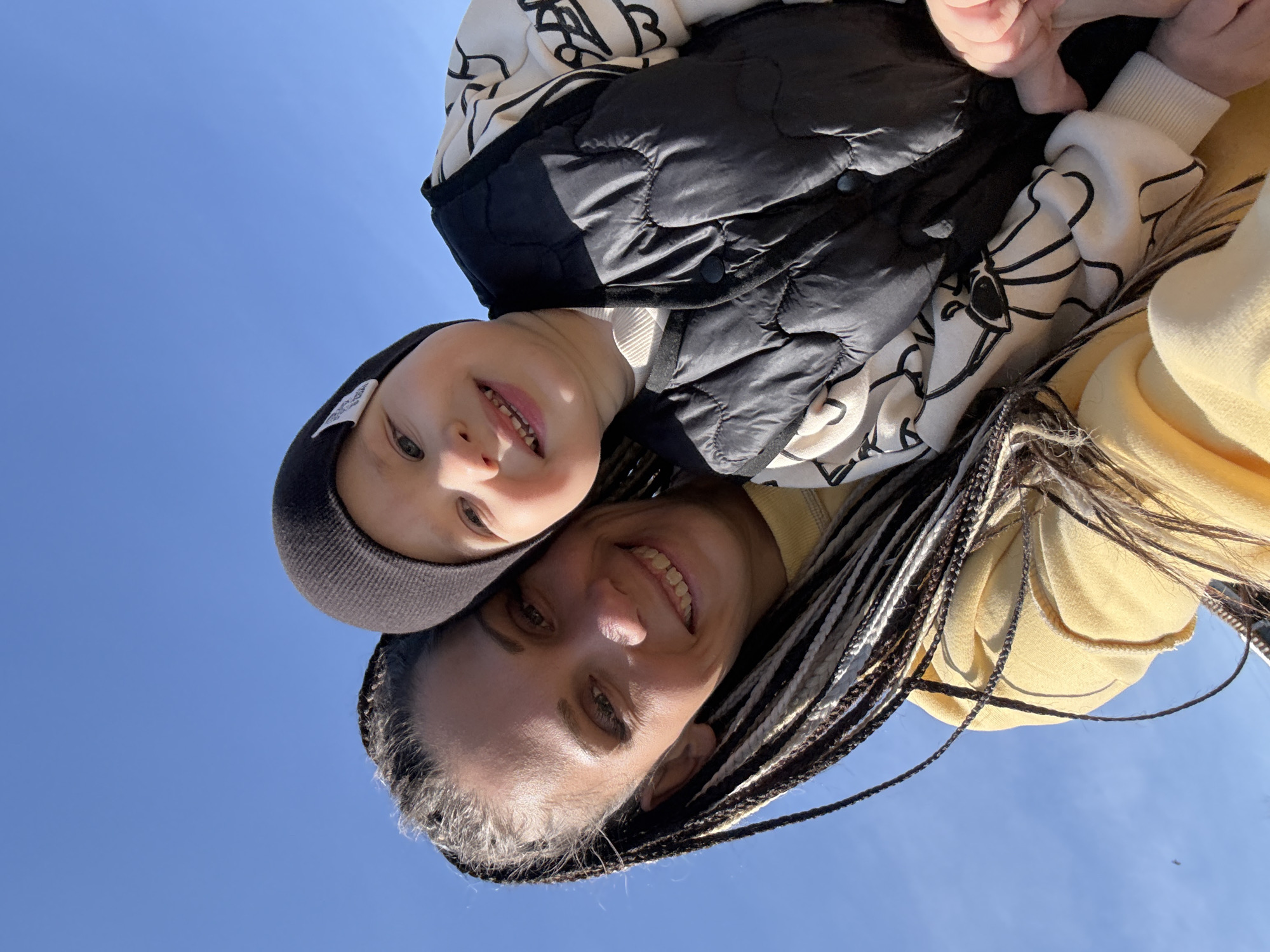 A selfie-style image taken from a low angle, looking up into a clear blue sky. A woman and a child are in the foreground, oriented upside down from the viewer's perspective. The woman is smiling and has long, braided hair. The child is wearing a knit cap, a patterned top, and a black quilted vest. They are positioned so that the child appears to be above the woman, possibly being held up or carried on her shoulders. The location is stated to be Rusanovo, Russia, but there are no specific environmental details in the image to confirm this.
